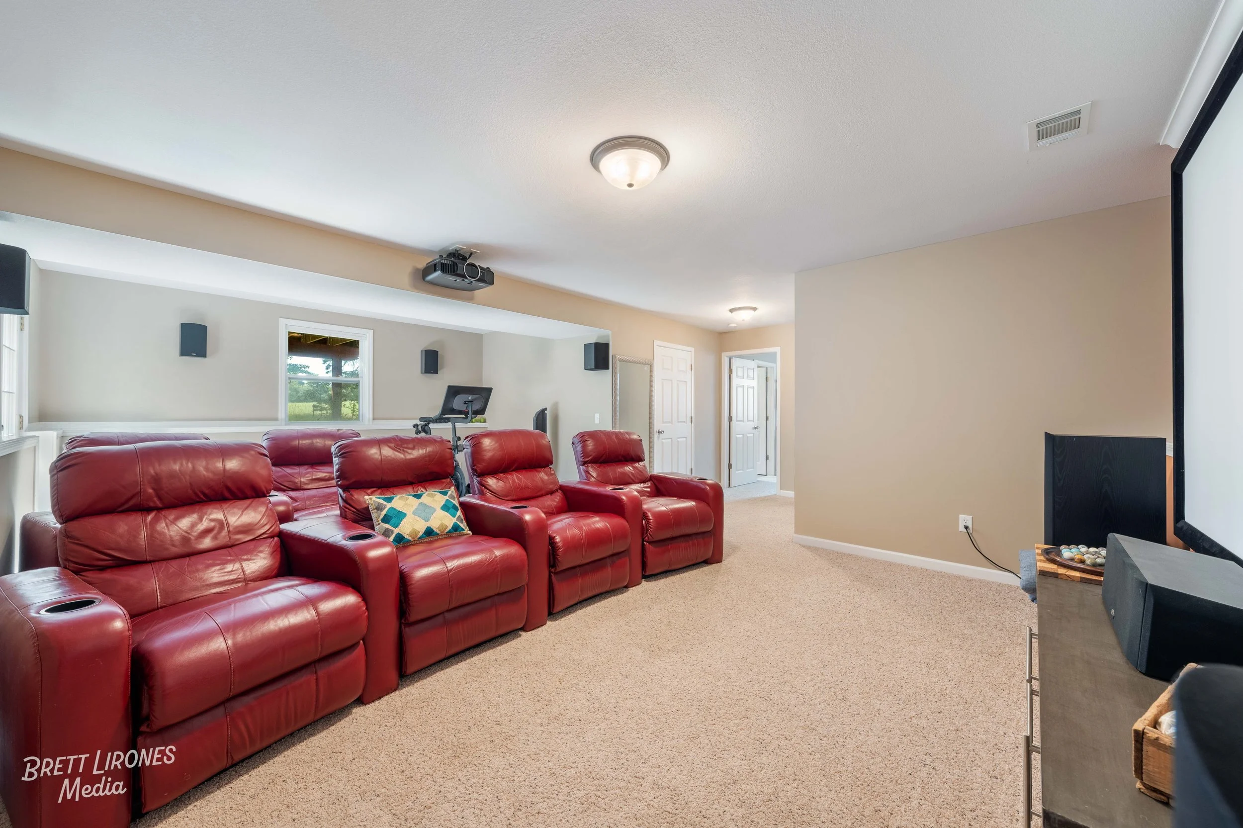 Living room with four red leather recliners, beige carpet, wall-mounted speakers, a projector, a large screen on the right, and a small table or cabinet with a speaker and decorative items.