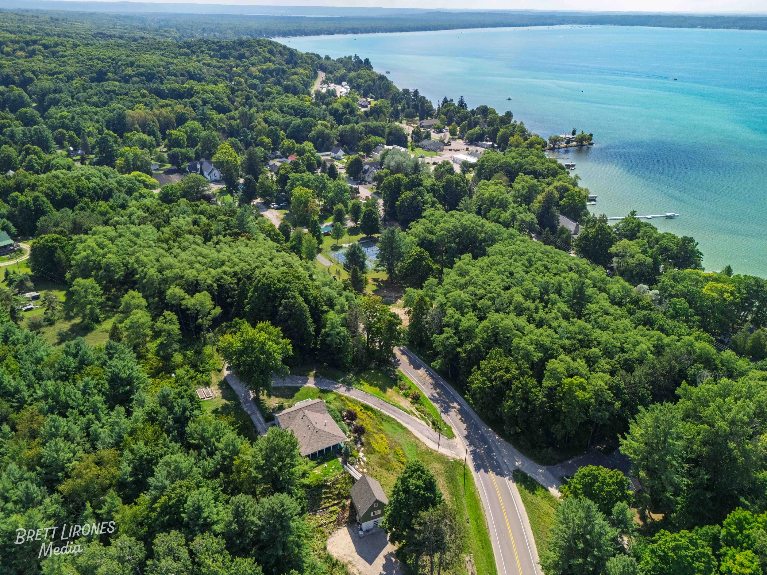 Aerial view of a lush green lakeside residential area with numerous trees, houses, and docks along the shoreline of a large blue lake, viewed from above on a sunny day.