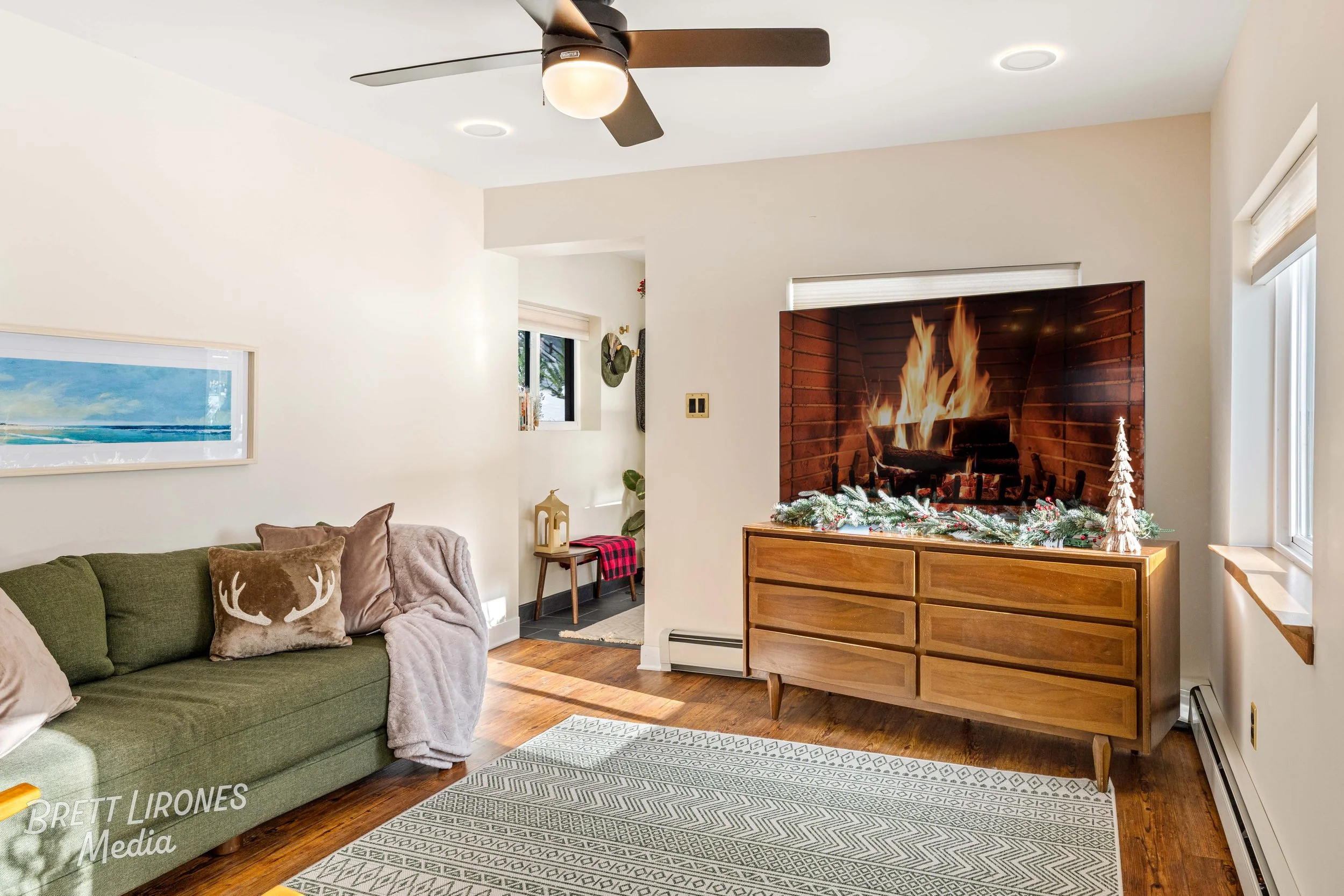 Living room with a green sofa, decorative pillows, and cozy throws, a wooden console with holiday decor, a fireplace displayed on the TV screen, hardwood flooring, a ceiling fan, and windows bringing natural light.