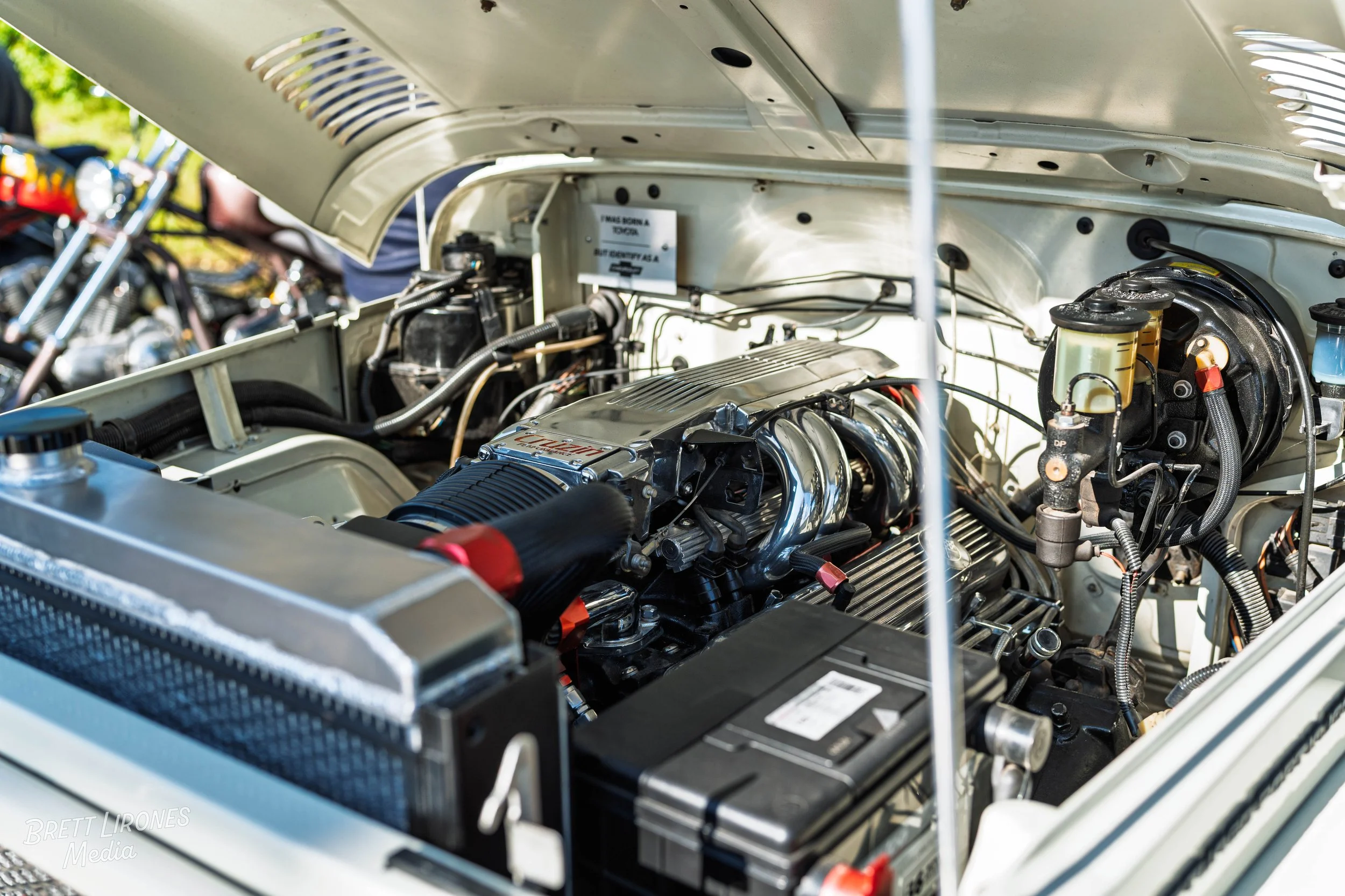 Close-up of a classic car engine with chrome components, black hoses, and metallic parts, with other motorcycles blurred in the background.