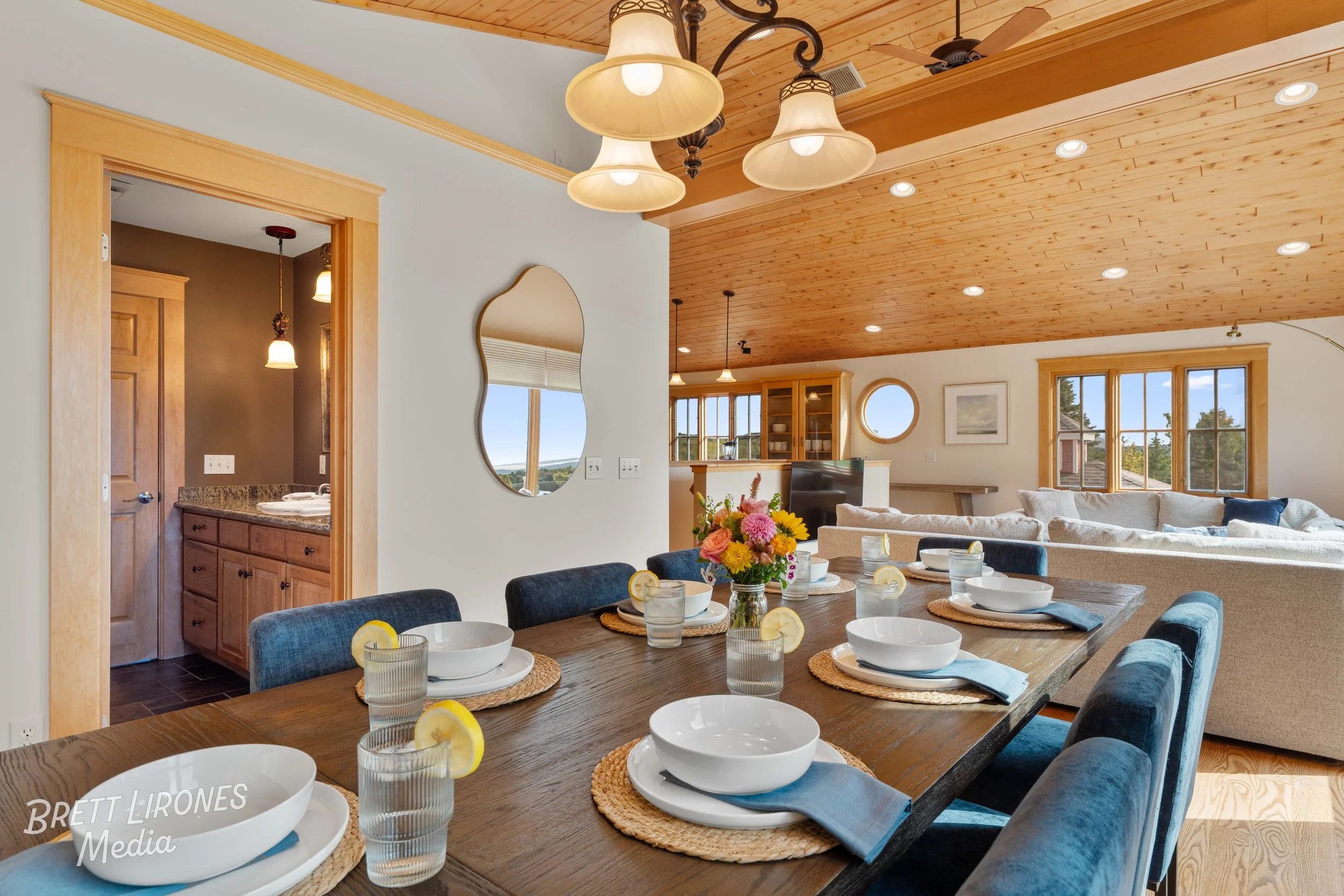 A dining table set for a meal with bowls, glasses with lemon slices, and a flower centerpiece, in an open-concept living room with wooden ceiling, large windows, and a cozy sofa.