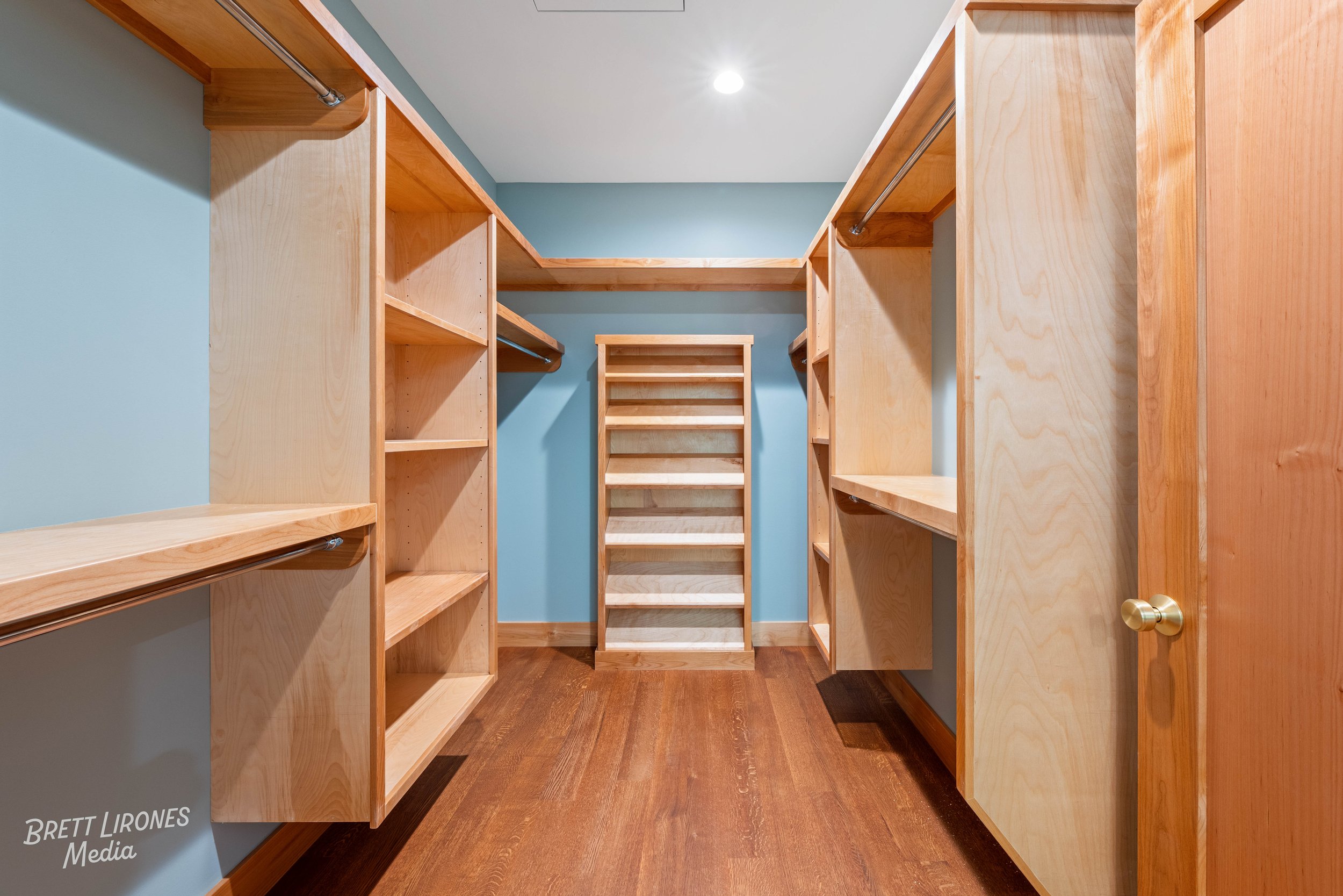Empty walk-in closet with wooden shelves, rods, and a central shelving unit, blue walls, and hardwood flooring.