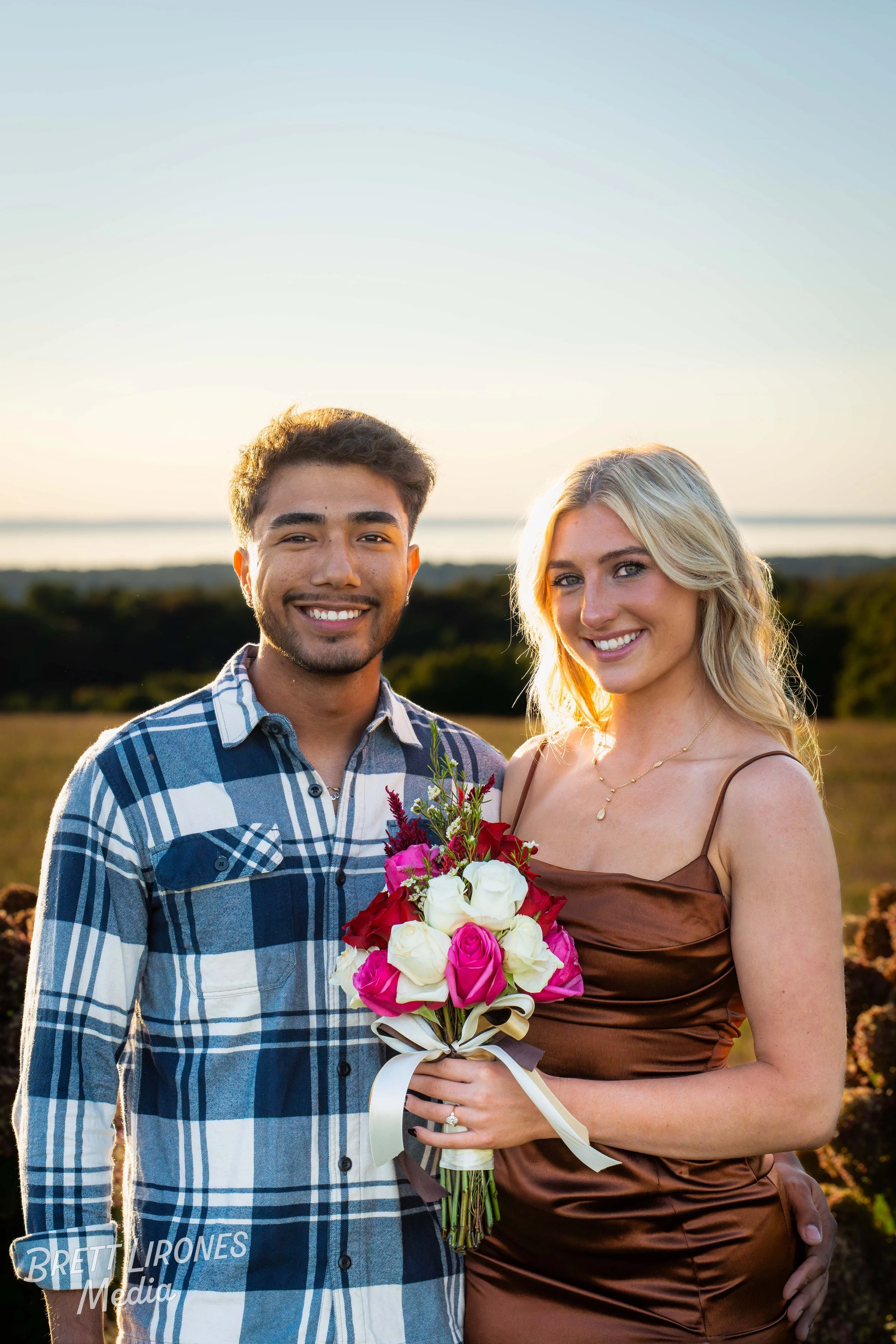 A smiling young man and woman standing outdoors at sunset, holding a bouquet of pink, white, and red roses.
