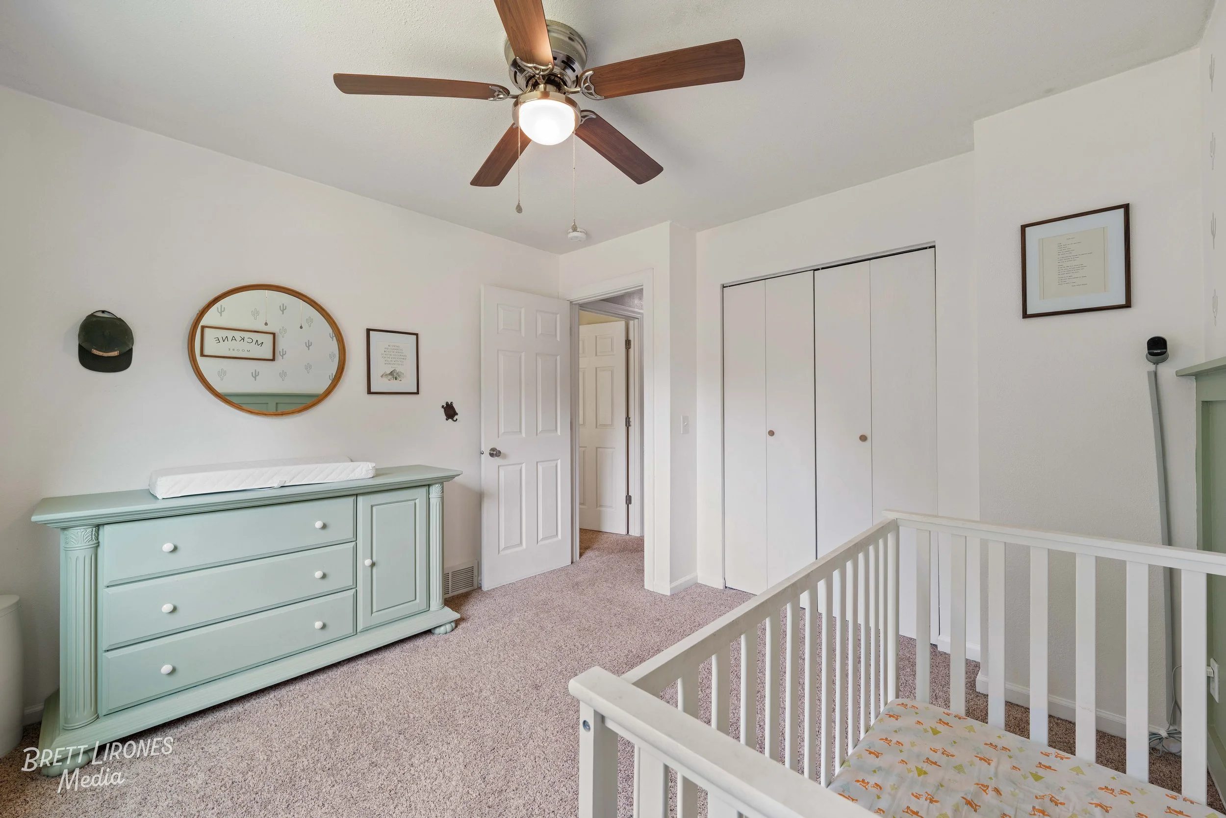 Bedroom with green dresser, mirror, wall art, ceiling fan, white crib, and closet doors.