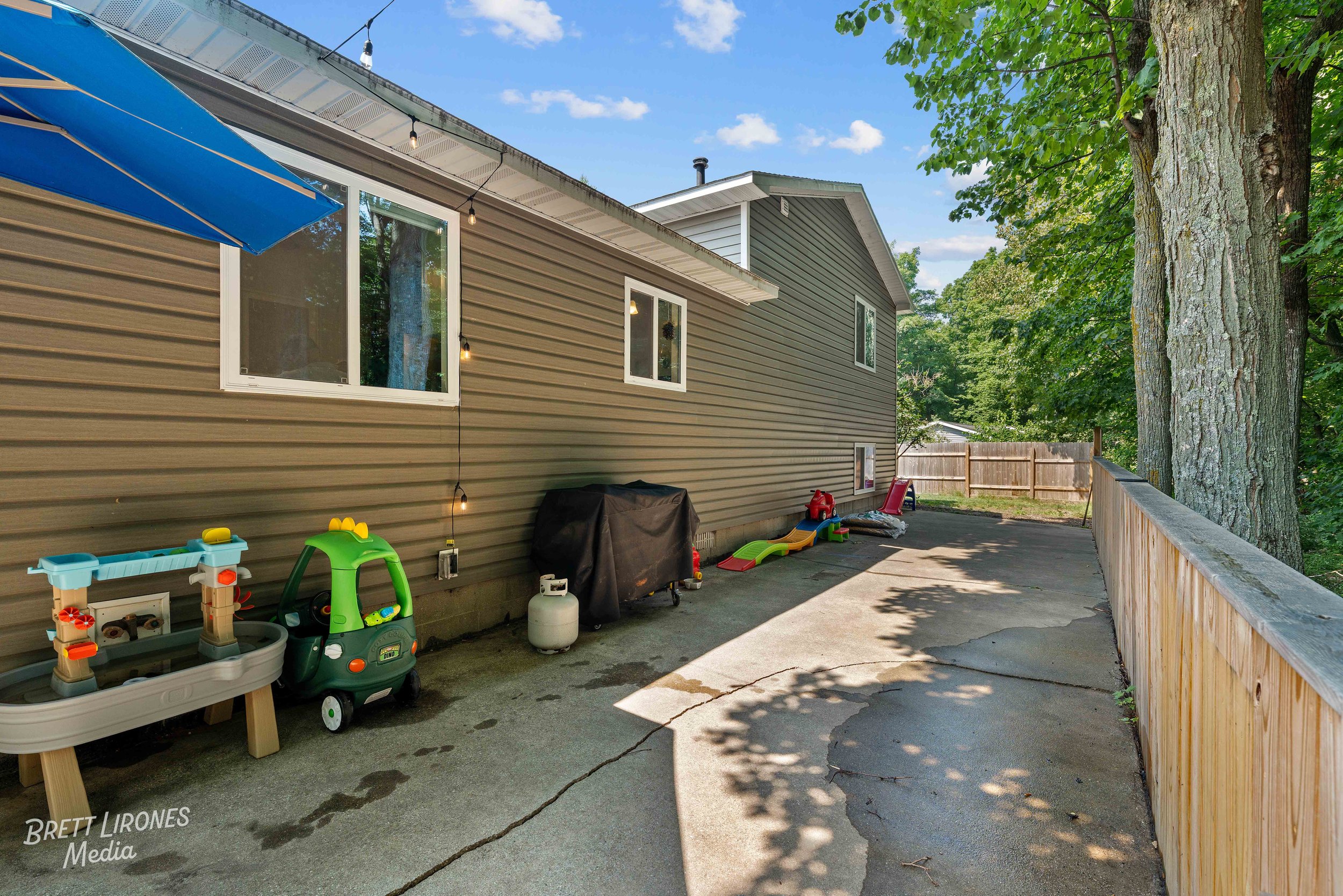 Back patio of a house with children's toys, a grill, and a wooden fence, with large trees and a blue sky in the background.