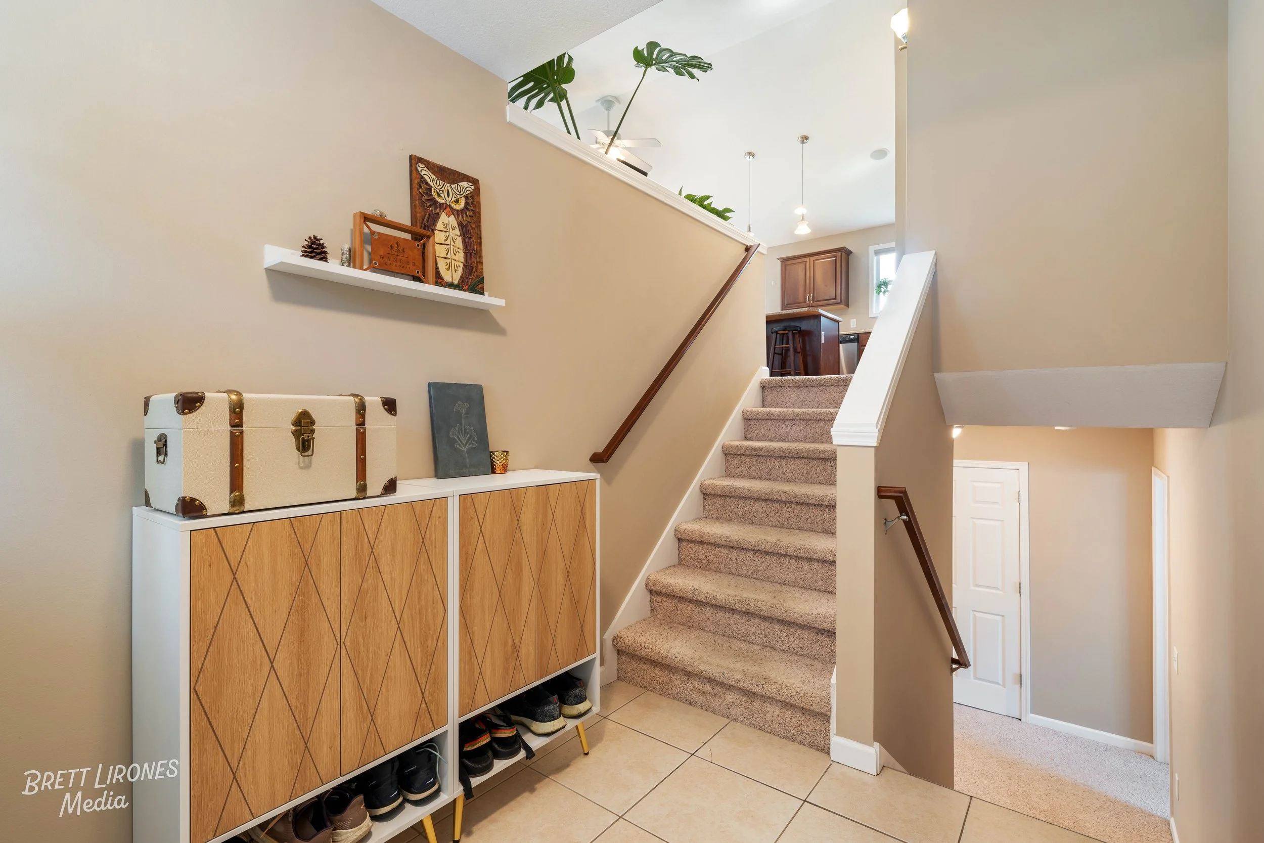 Entryway of a home with beige walls, a staircase with carpeted steps, a wooden handrail, and a white and wood console table with storage and shoes underneath. Decorations include framed art, a small canvas, a vintage suitcase, and a wall shelf with d