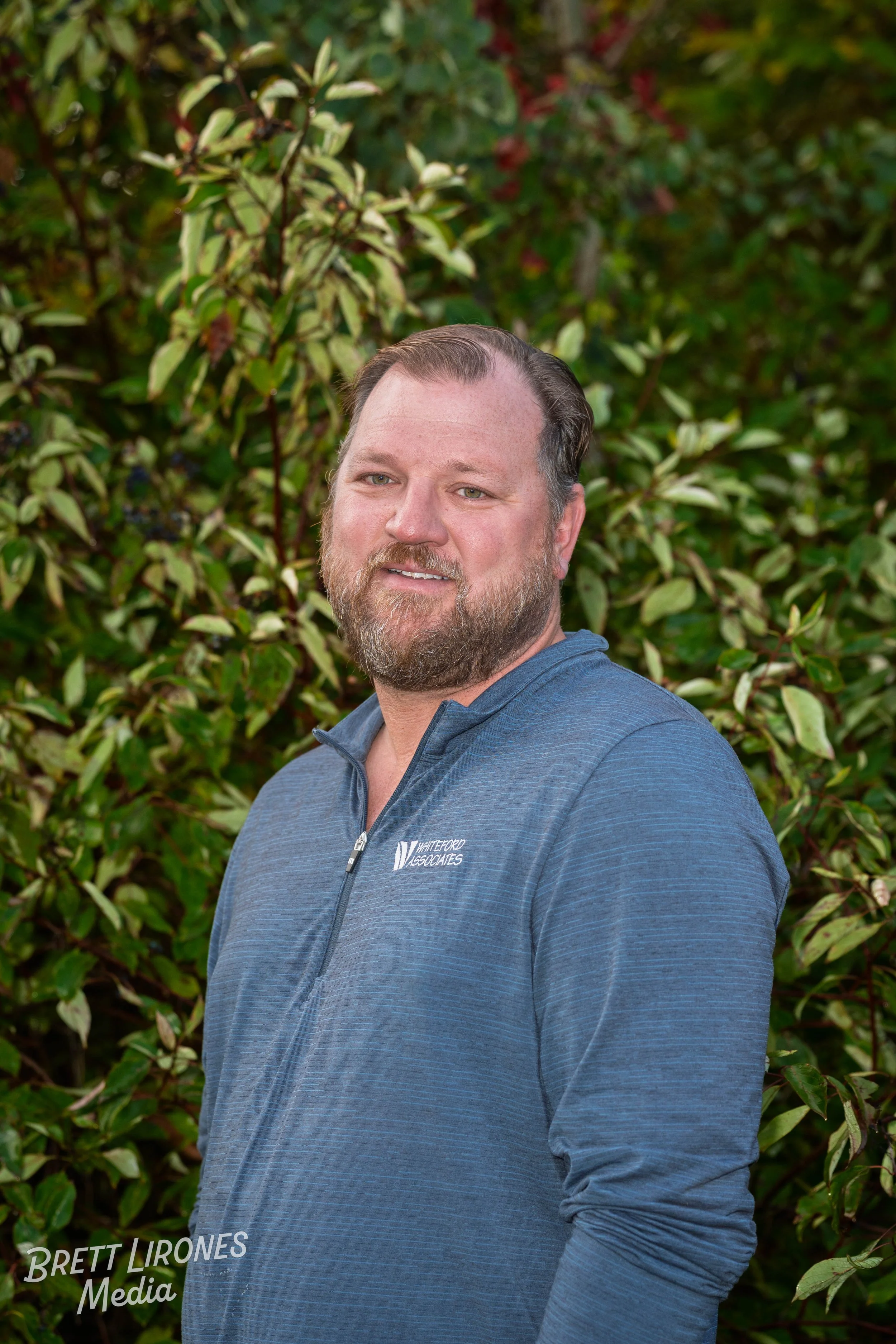 A man with a beard and short hair wearing a blue quarter-zip jacket standing outdoors in front of leafy bushes.