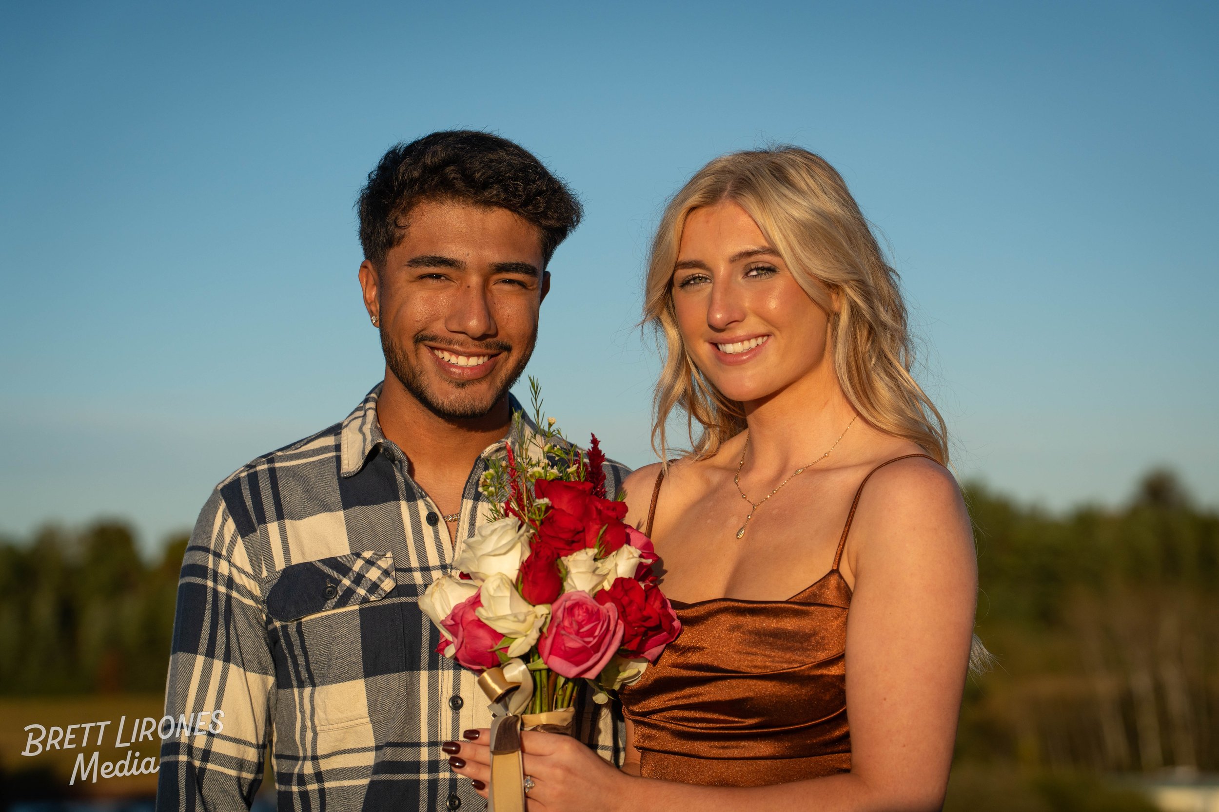 A happy couple, a man and a woman, smiling outdoors with a clear blue sky in the background. The woman is holding a bouquet of pink, white, and red roses.