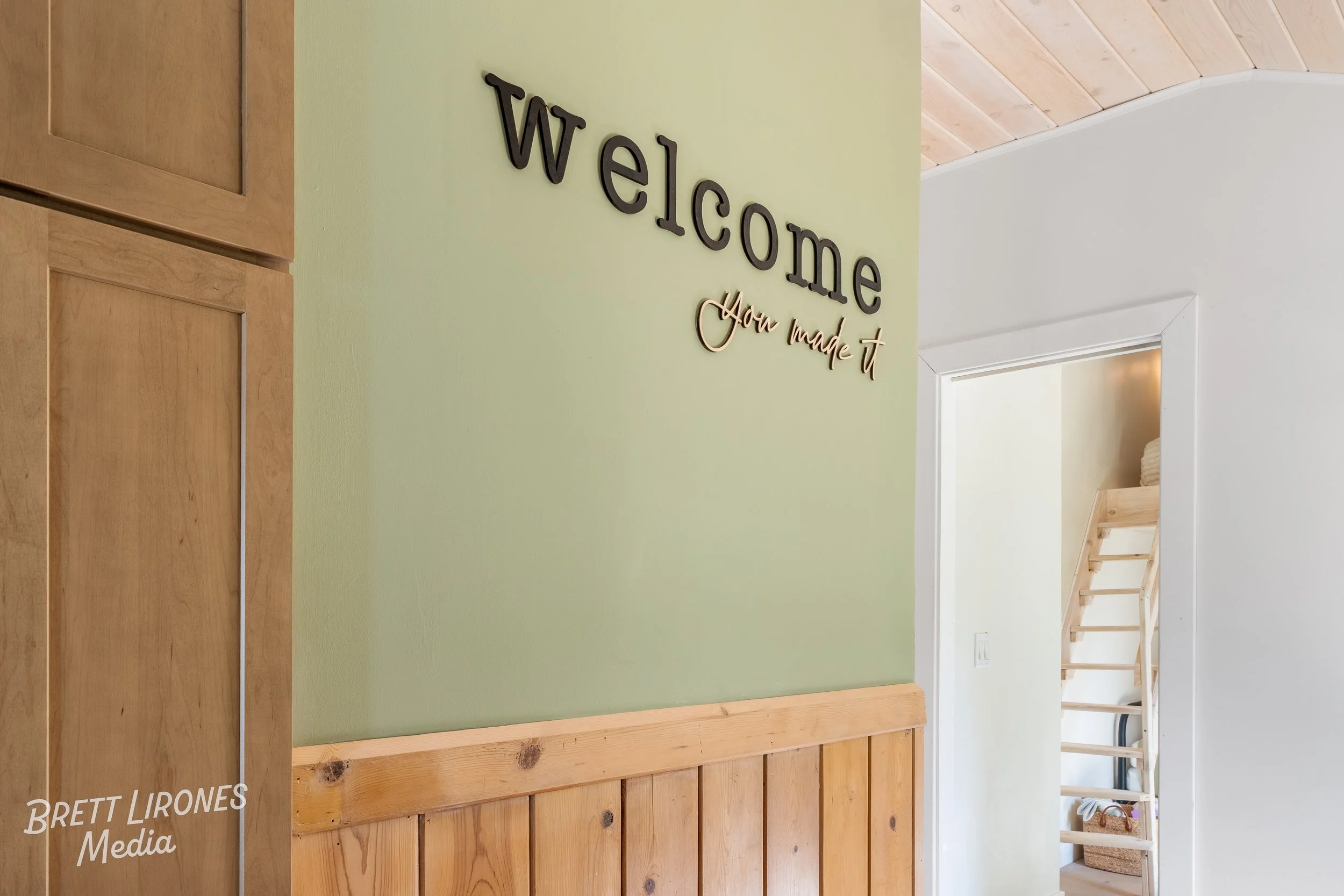 A light green wall with black and gold decorative sign saying 'welcome you made it' in a home interior. Part of a wooden cabinet or wall panel is visible on the left, and a doorway leads to a staircase in the background.