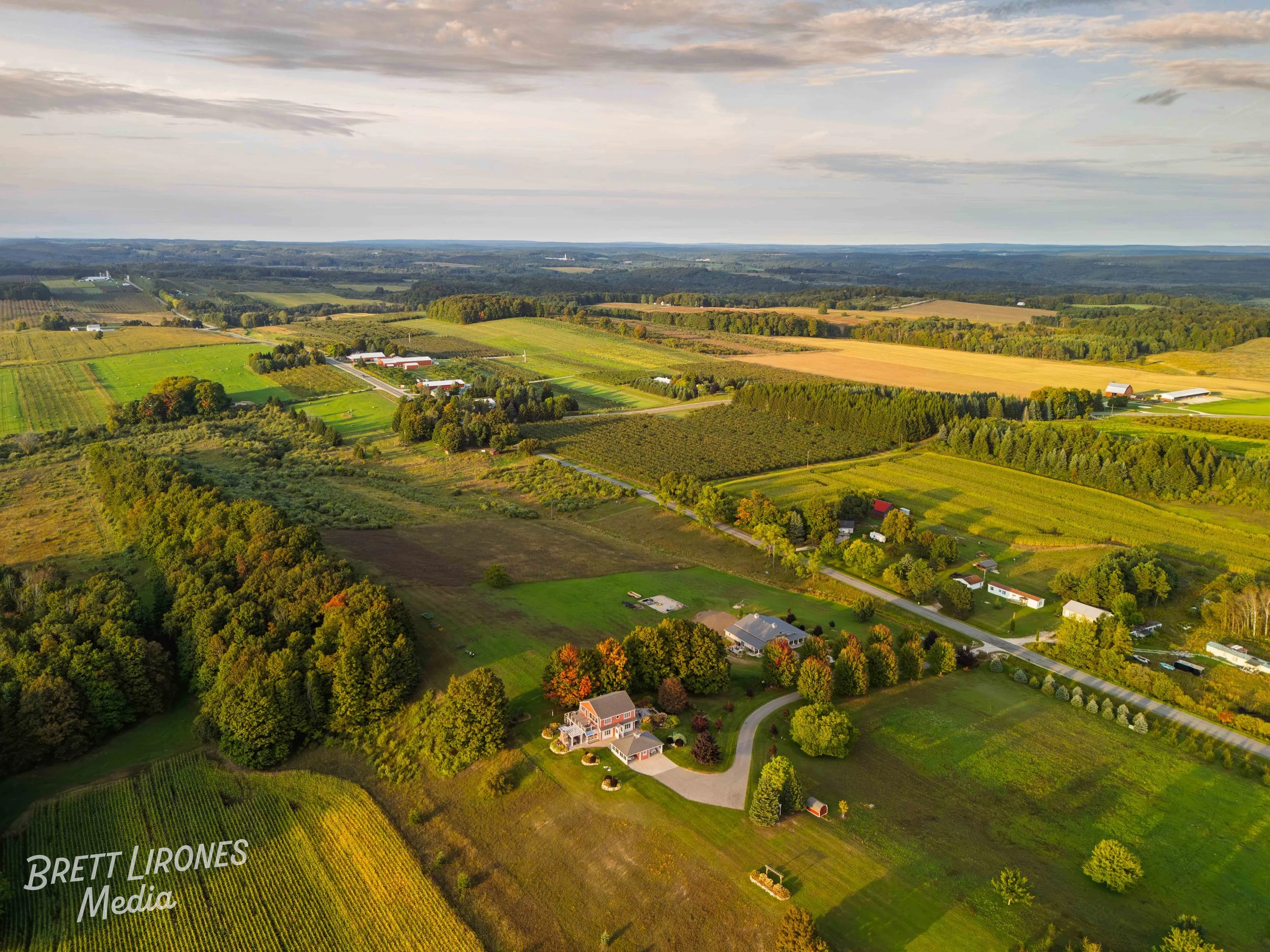 Aerial view of a rural landscape with farmland, trees, and houses under a partly cloudy sky during sunset.