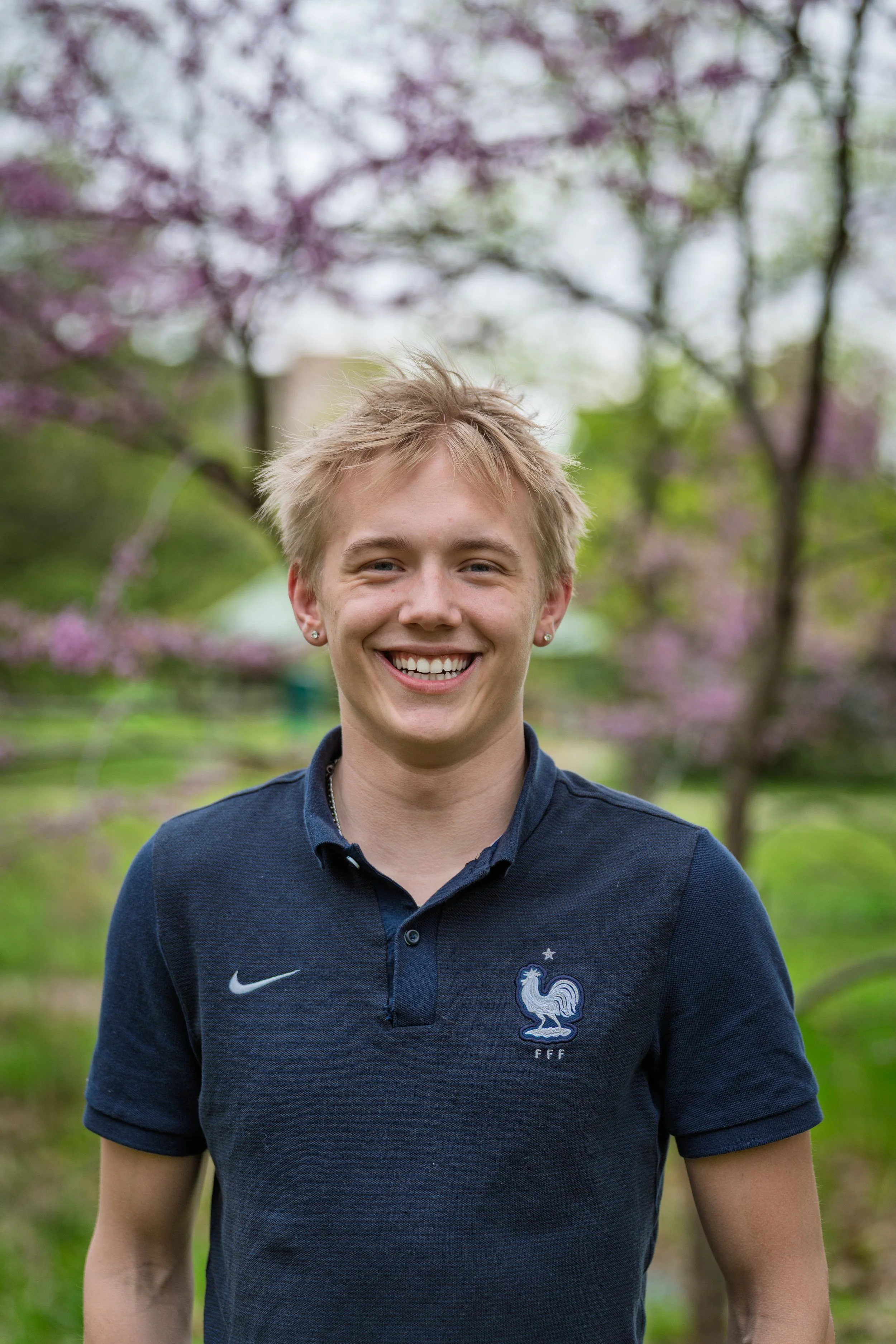 Young man smiling outdoors, wearing a navy blue polo shirt with the French national football team logo, with a background of spring trees with pink blossoms.