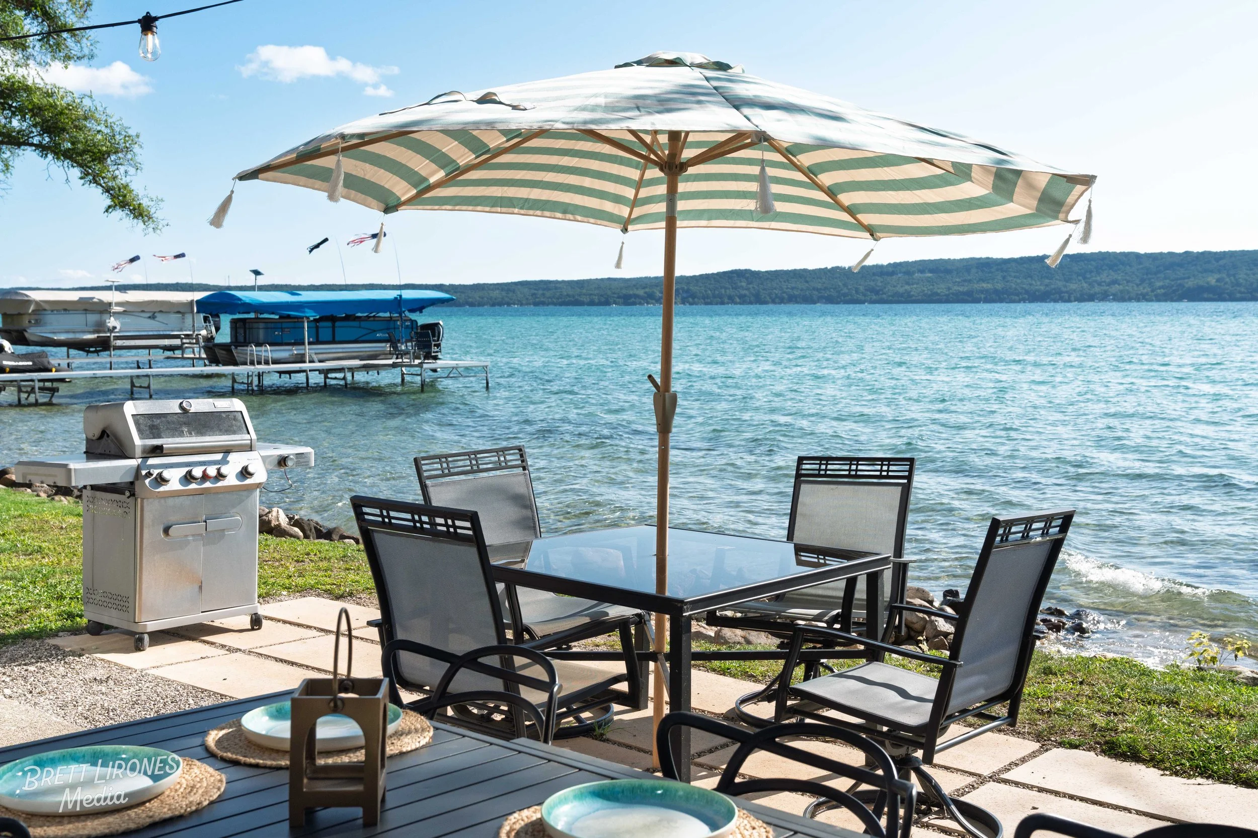 A lakeside outdoor patio with a striped umbrella, black metal dining table and chairs, a barbecue grill, and a view of the lake with boats and a dock in the background.