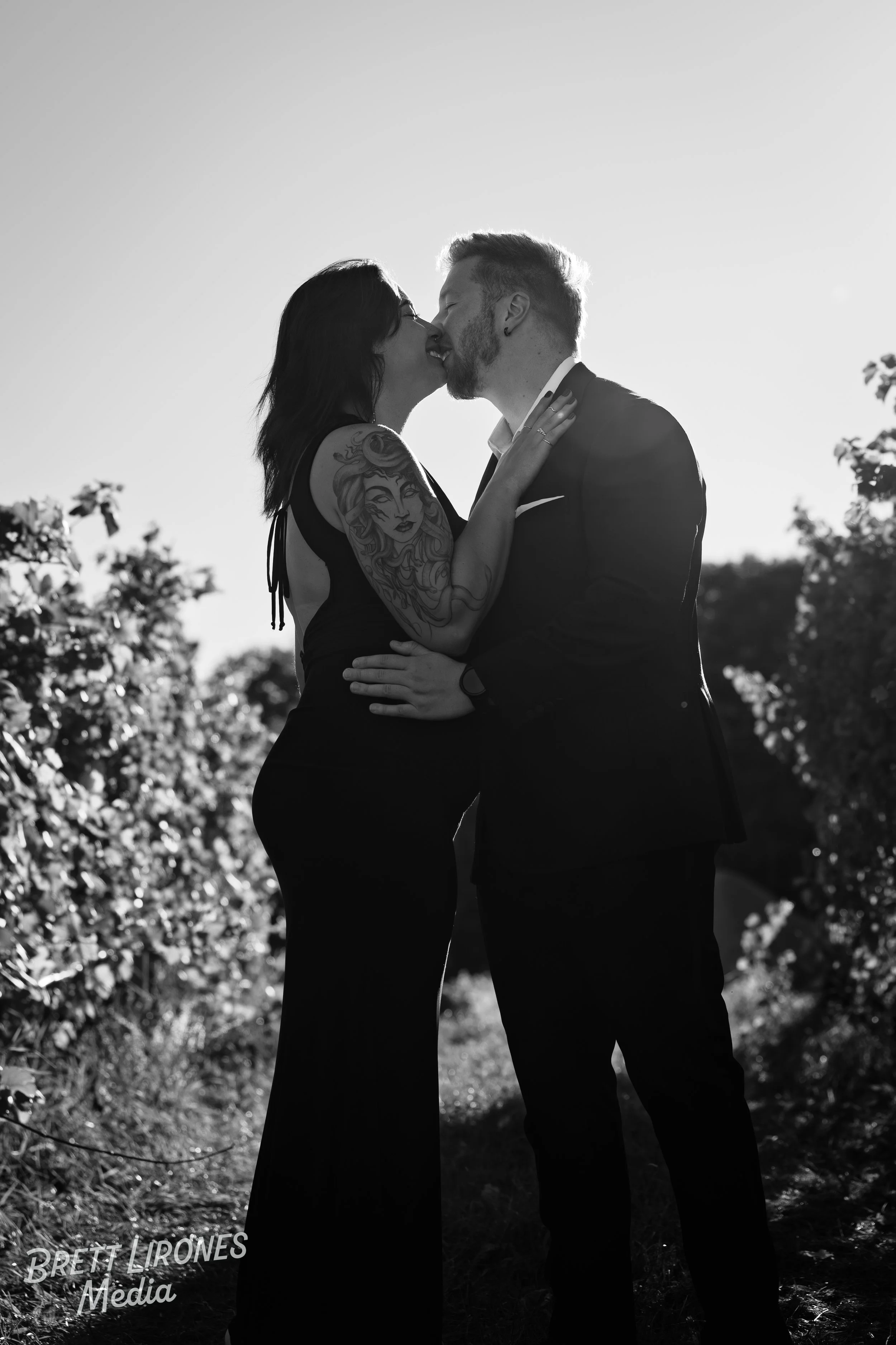 A couple sharing a kiss outdoors, the woman with tattoos on her arm and shoulder, and the man in a suit, backlit by the sun, outdoors with trees in the background.
