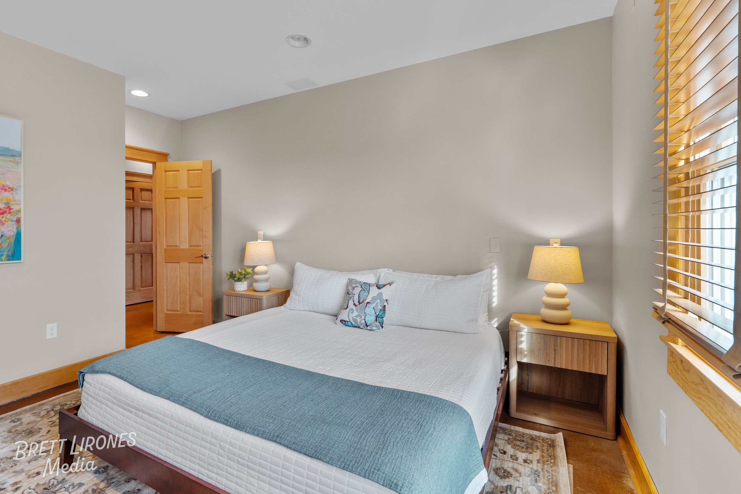 A bedroom with a bed, two nightstands with lamps, a window with blinds, and a partially open wooden door.