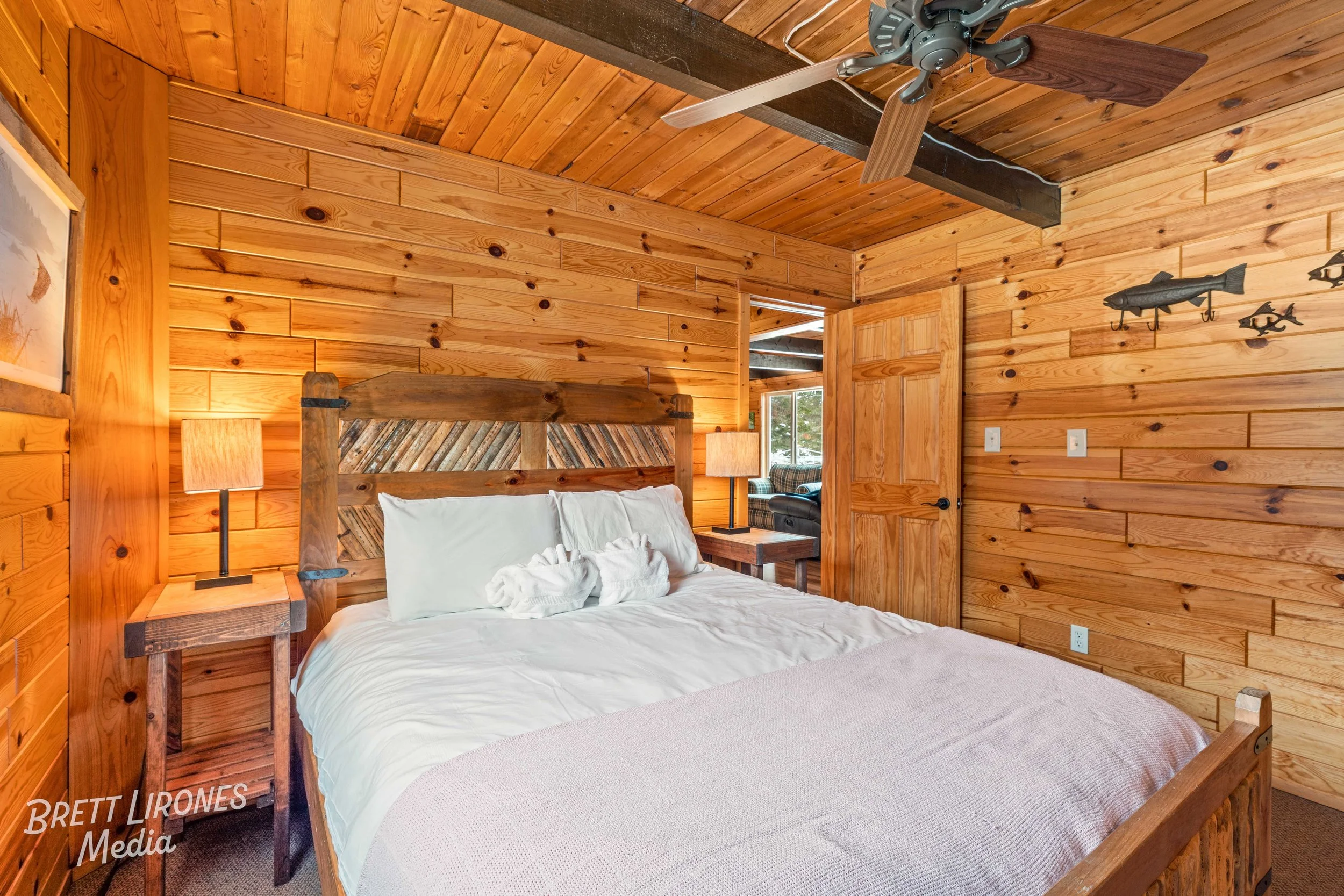 A cozy bedroom with wooden walls, featuring a bed with white bedding and an artistic headboard. There are two matching nightstands each with a lamp, and a ceiling fan. A door to another room is open, showing a glimpse of a living area with a couch an