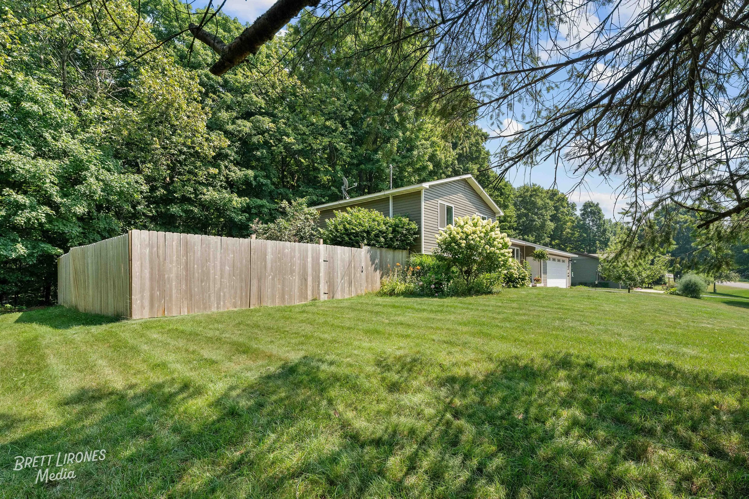 A backyard with a green lawn, a wooden fence, a gray house with a garage, surrounded by trees and bushes under a partly cloudy sky.