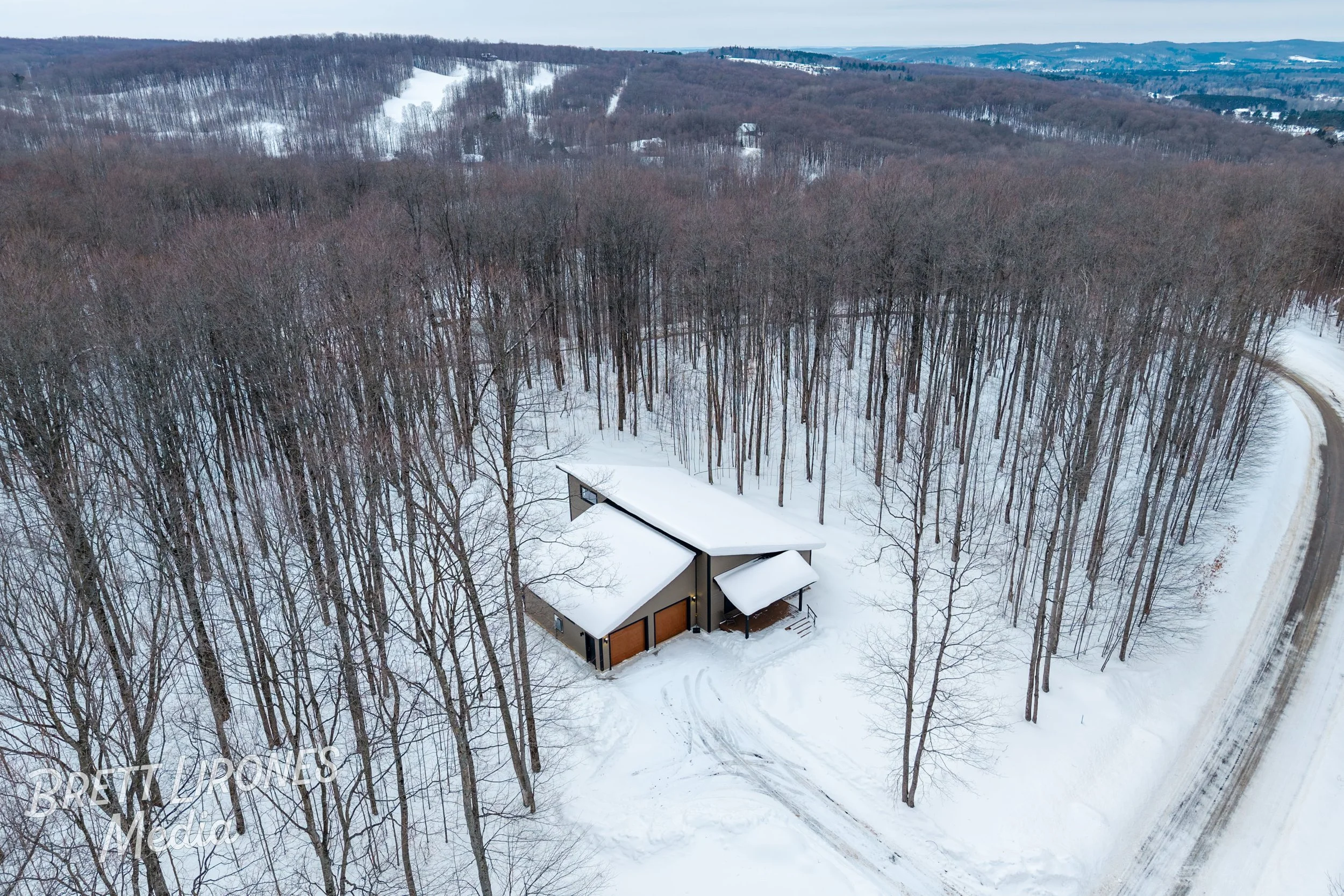 A house surrounded by leafless trees during winter with snow on the ground and on the house's roof, situated near a winding road.