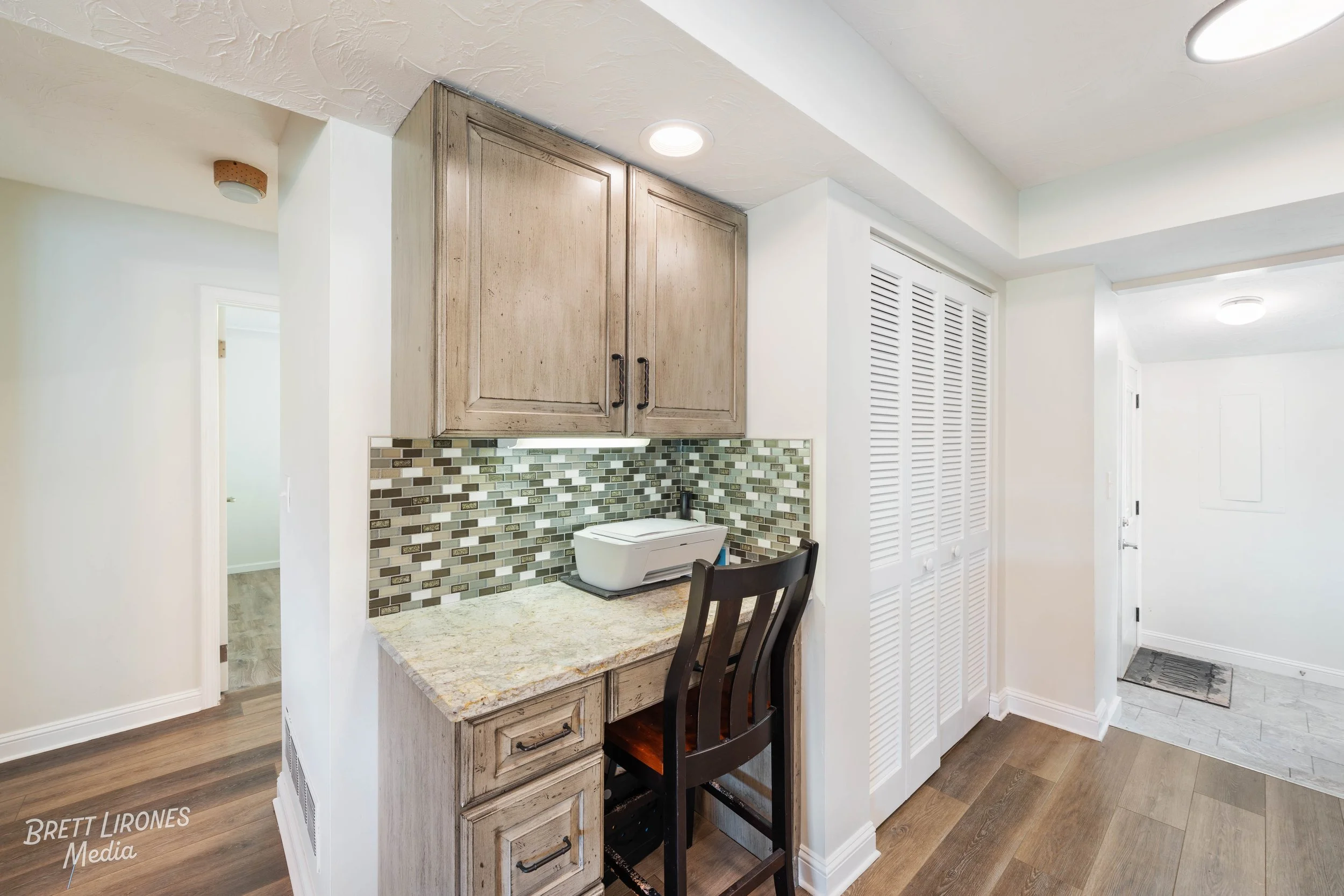A small kitchen or pantry area with light-colored wooden cabinets, a granite countertop, and a mosaic tile backsplash. A black chair is positioned at the counter, and there are white louvered closet doors nearby. The space has recessed ceiling lights