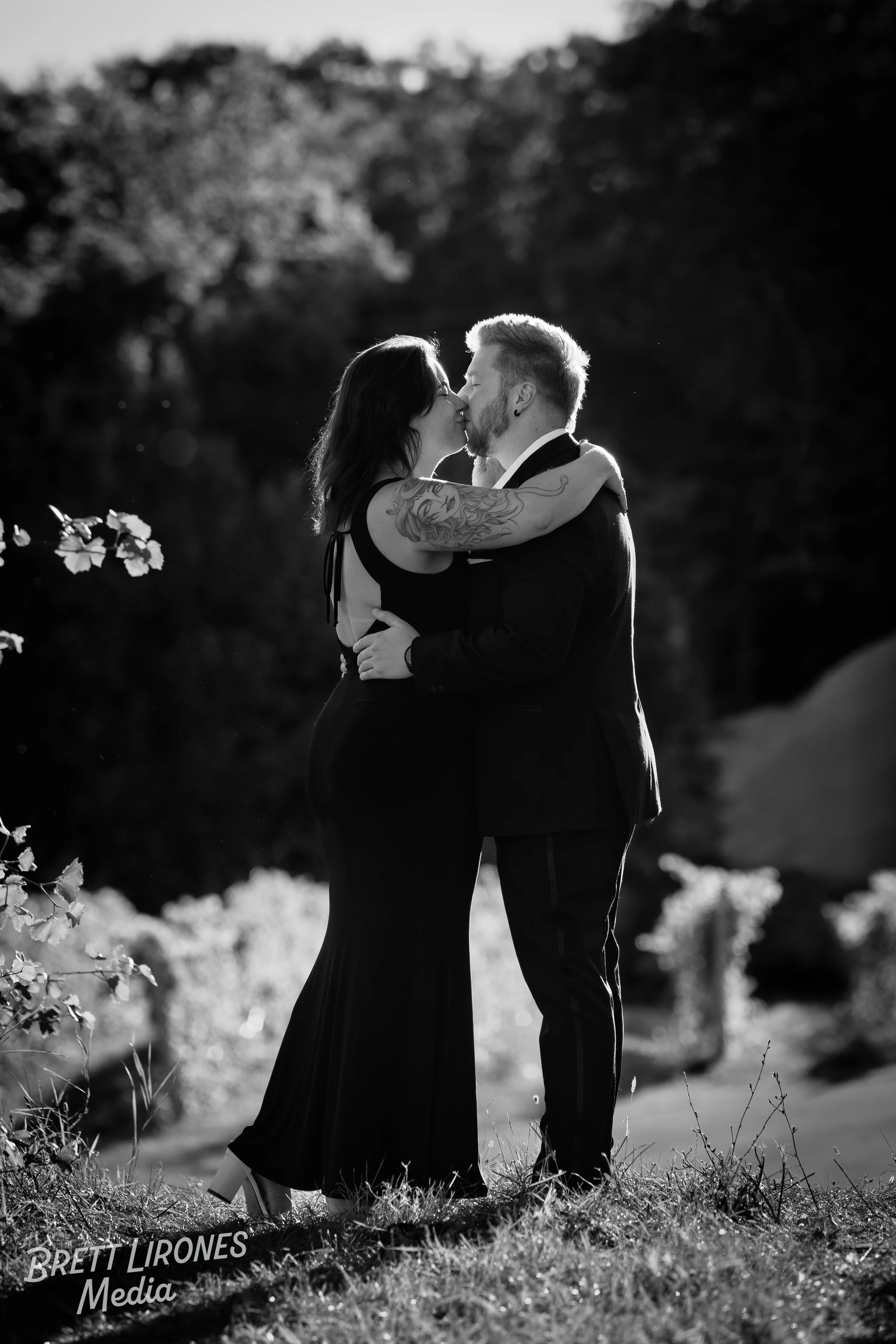 A black-and-white photograph of a couple kissing outdoors, with the woman wearing a long dress and the man in a suit. They are embracing on a grassy area with trees in the background, and the woman has tattoos on her arm.
