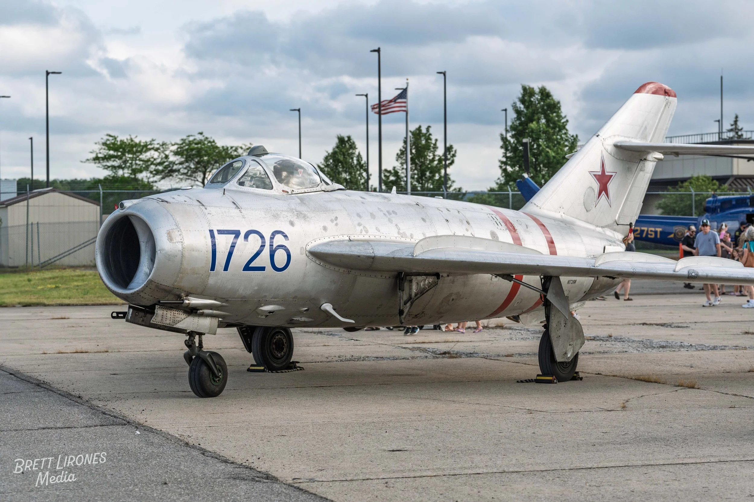 Military jet aircraft with the number 1726 and a red star insignia on its tail, parked on an airfield with people and trees in the background.