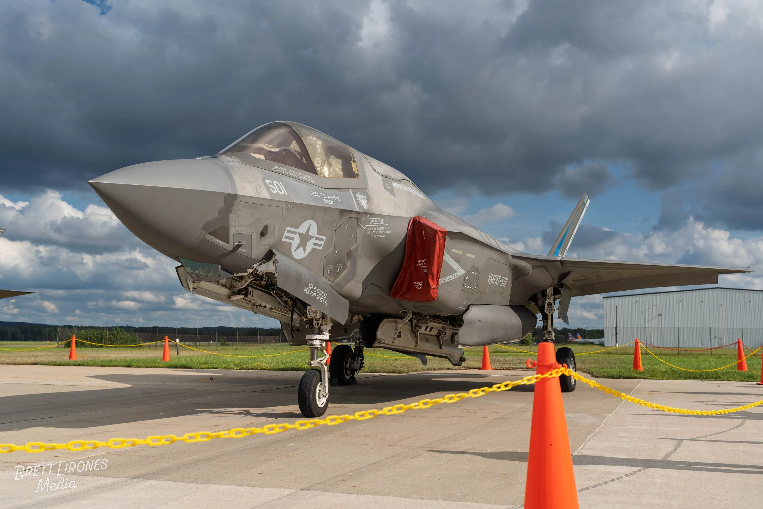 F-35 fighter jet parked outdoors on tarmac, surrounded by orange safety cones and chain barriers, under a cloudy sky.