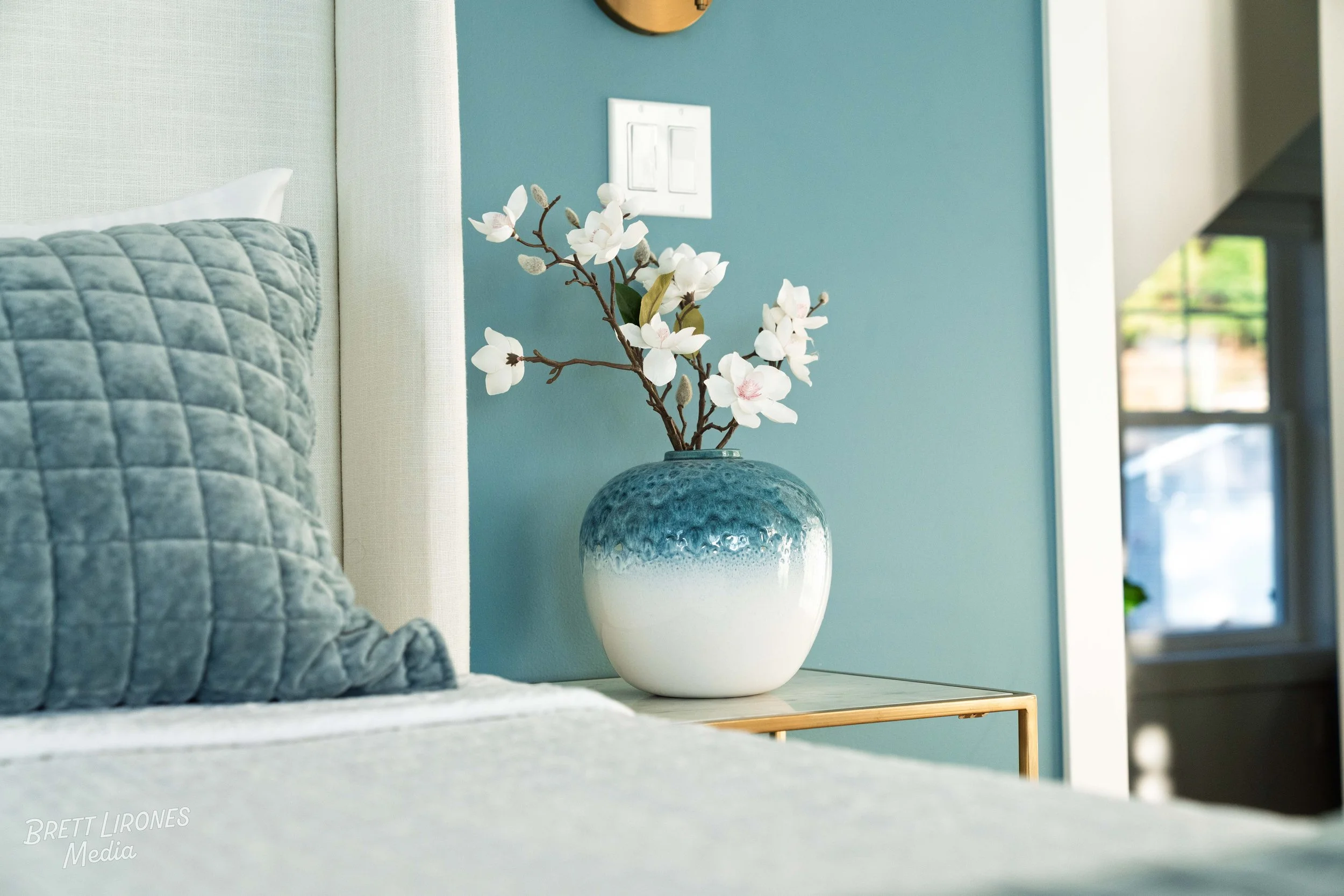 A bedroom nightstand with a ceramic vase holding white magnolia flowers, next to a bed with a quilted gray pillow, against a teal wall with a white light switch and a small window in the background.