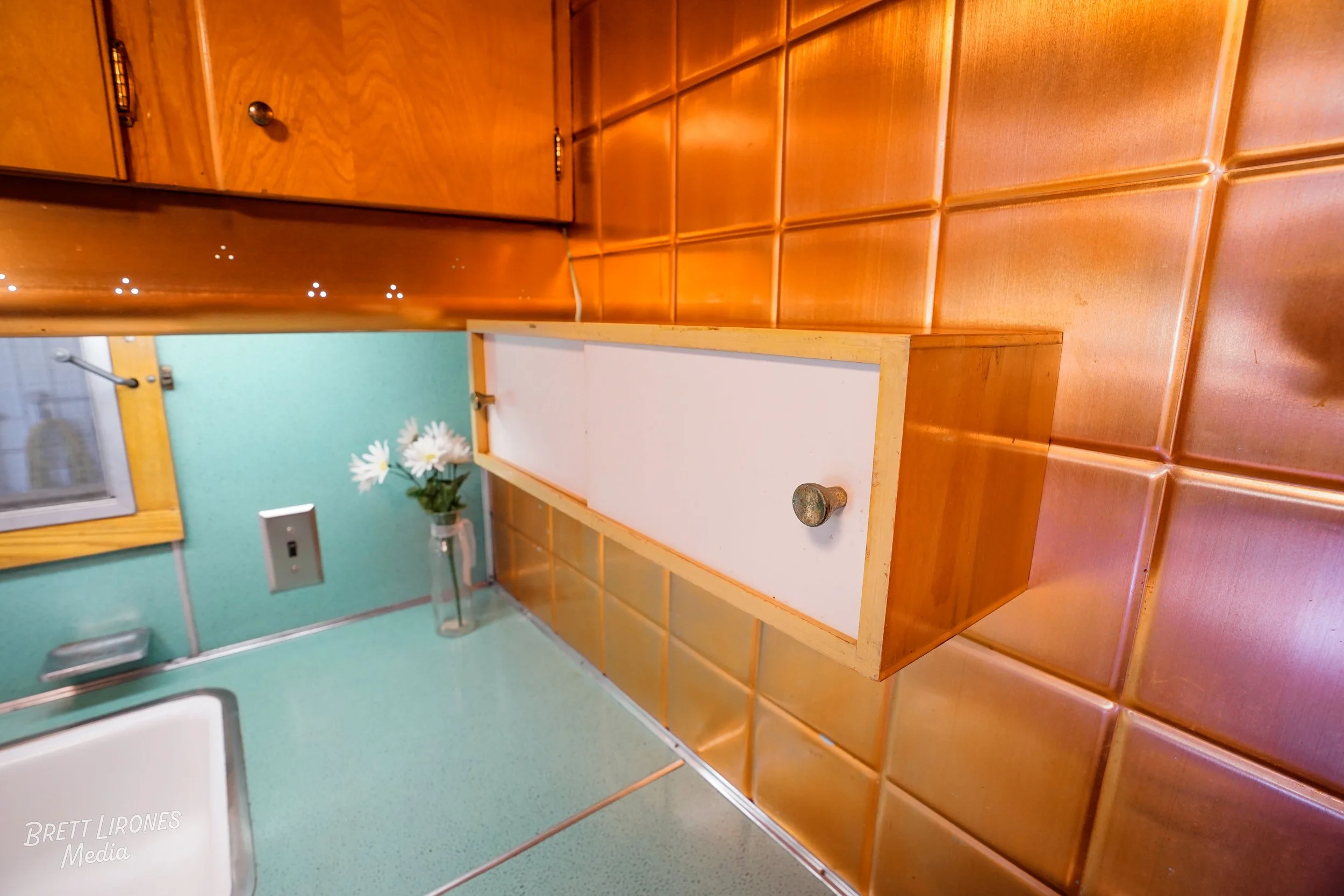 Kitchen corner with green countertop, small white cabinet with a knob, faucet, orange tile backsplash, and a window with a wooden frame, decorated with a vase of white daisies.
