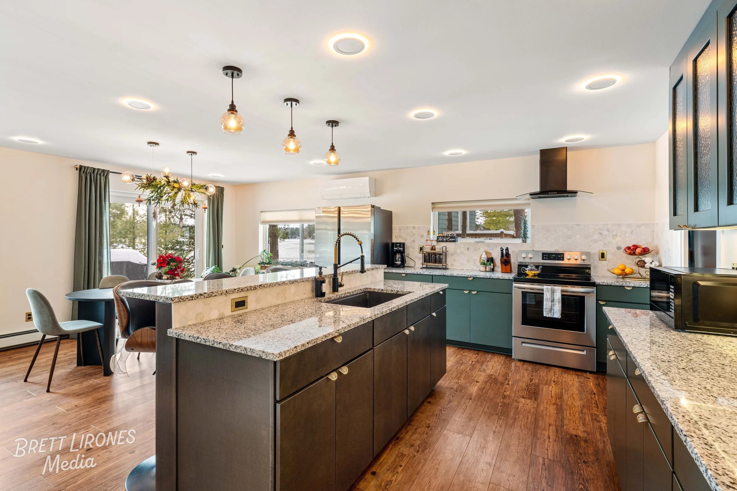 Modern kitchen with granite countertops, teal and dark brown cabinets, stainless steel appliances, and hardwood floors. There is a central island with a sink, a dining table with chairs, and a window with curtains overlooking trees.