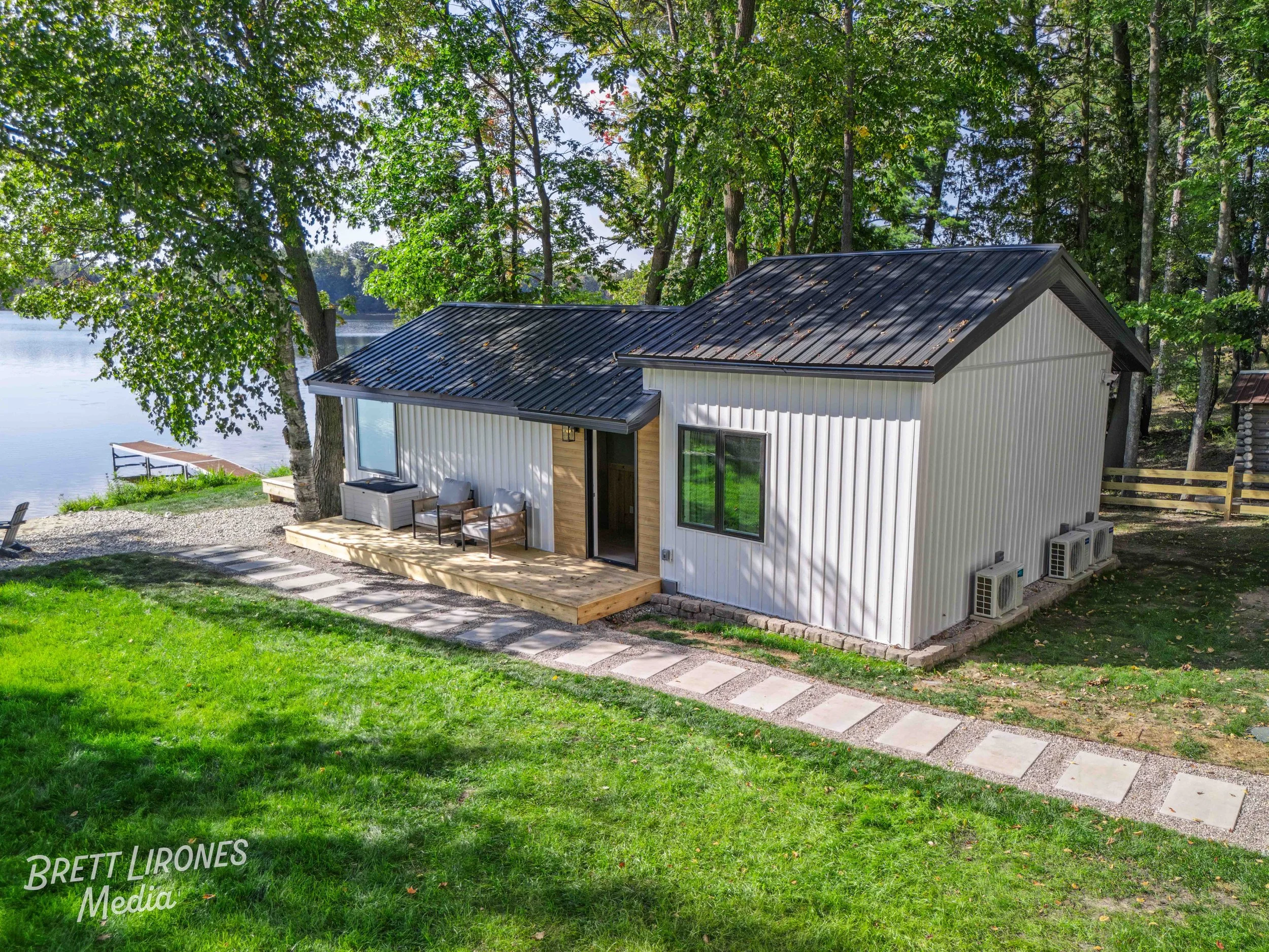 A small white house with a black metal roof is situated near a lakeside surrounded by trees, with a wooden deck and outdoor seating, overlooking the water.