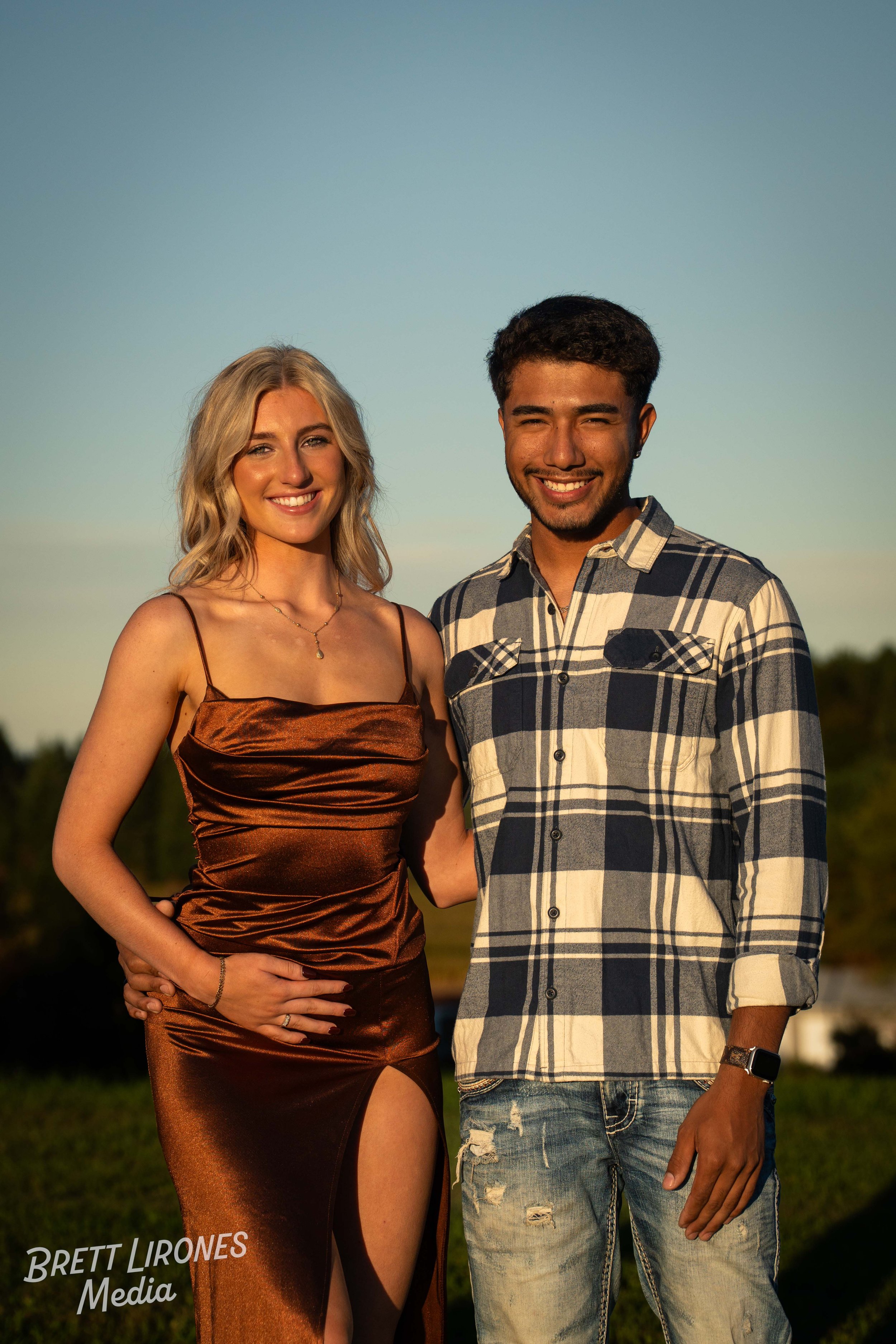 A young woman in a brown satin dress with a slit and spaghetti straps and a young man in a plaid shirt with rolled-up sleeves and ripped jeans, both smiling outside during sunset.