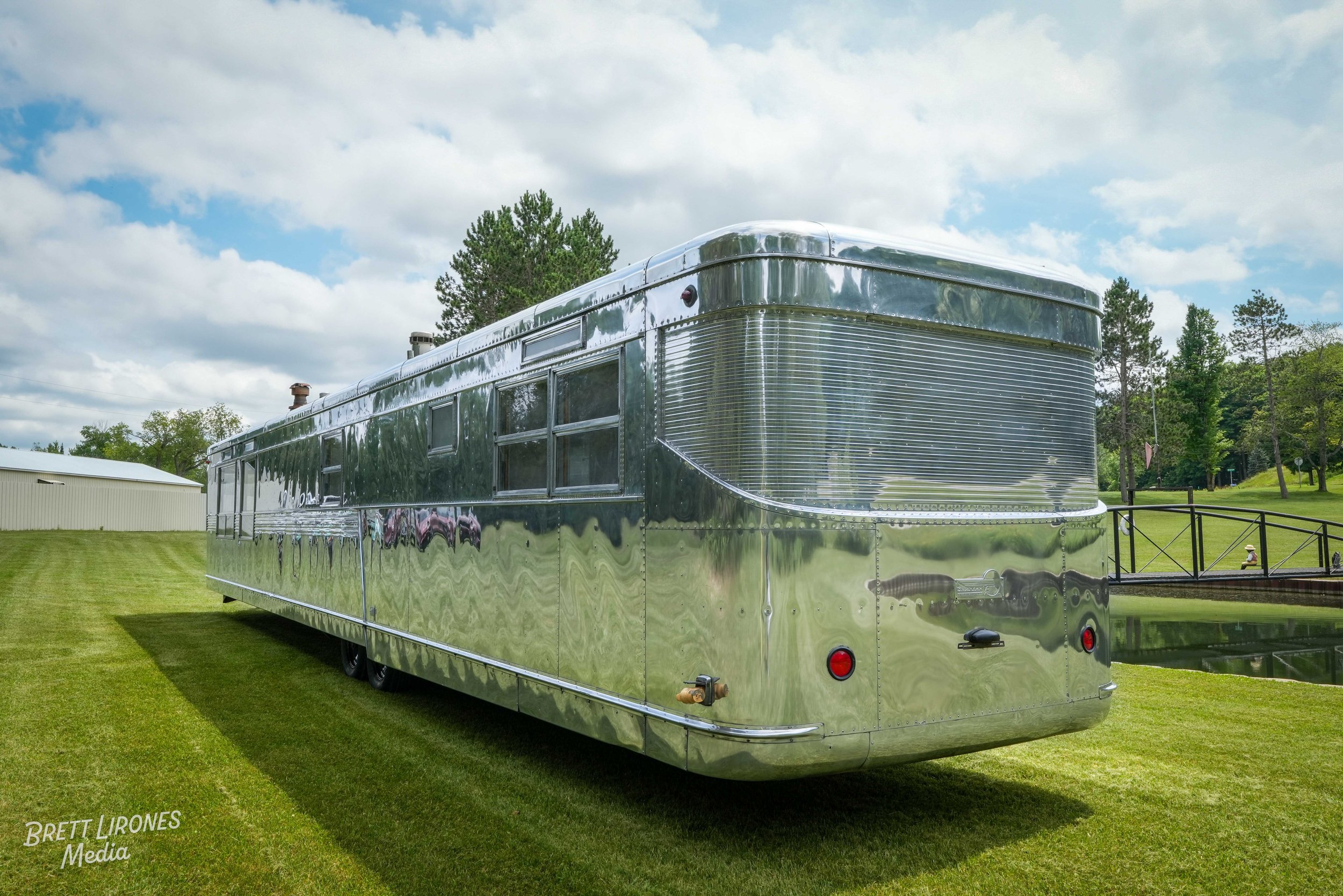 A shiny, vintage trailer with a metallic exterior parked on a grassy area with trees and a cloudy sky in the background.