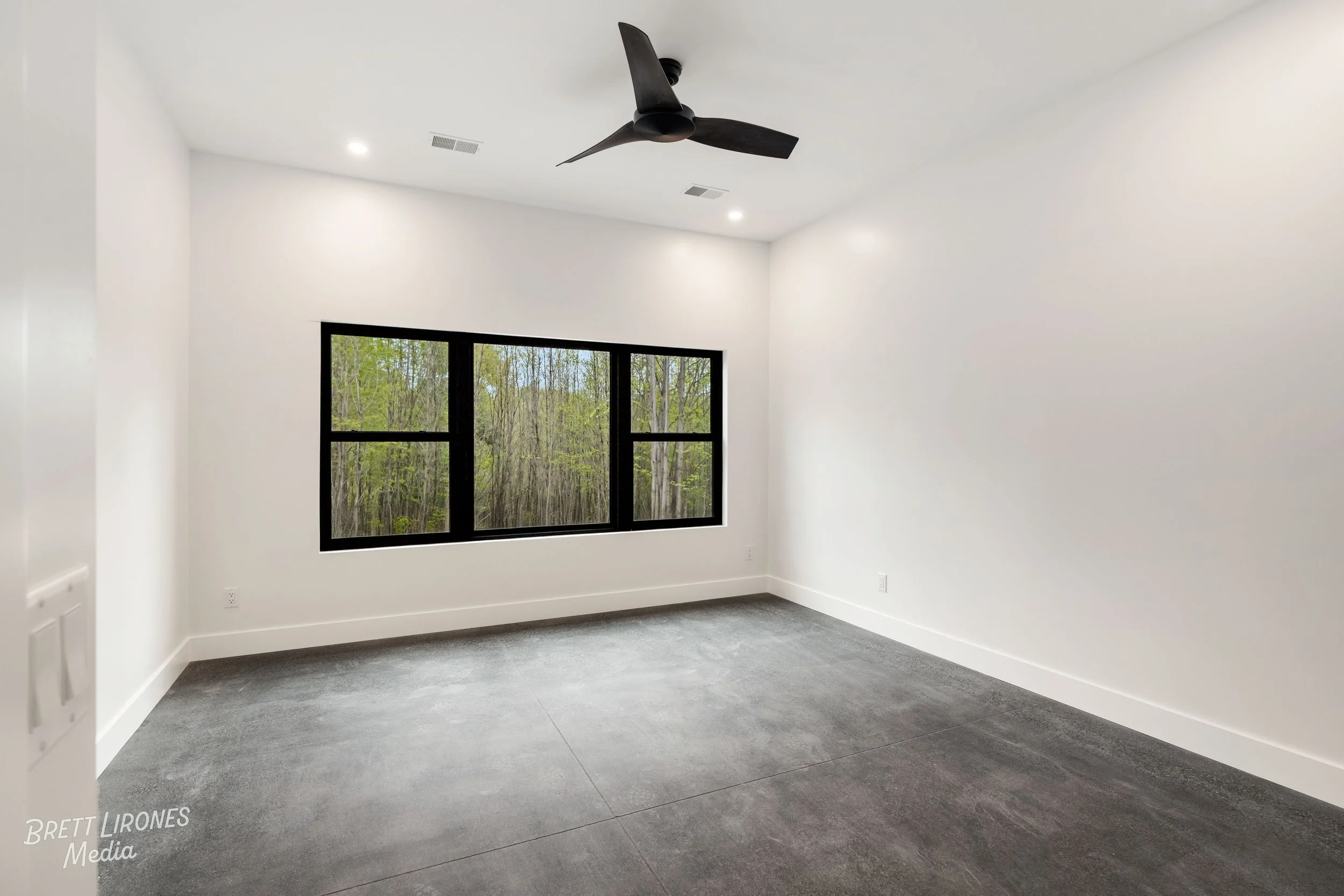 Empty white room with large window showing trees outside, gray flooring, black ceiling fan, and recessed lights.