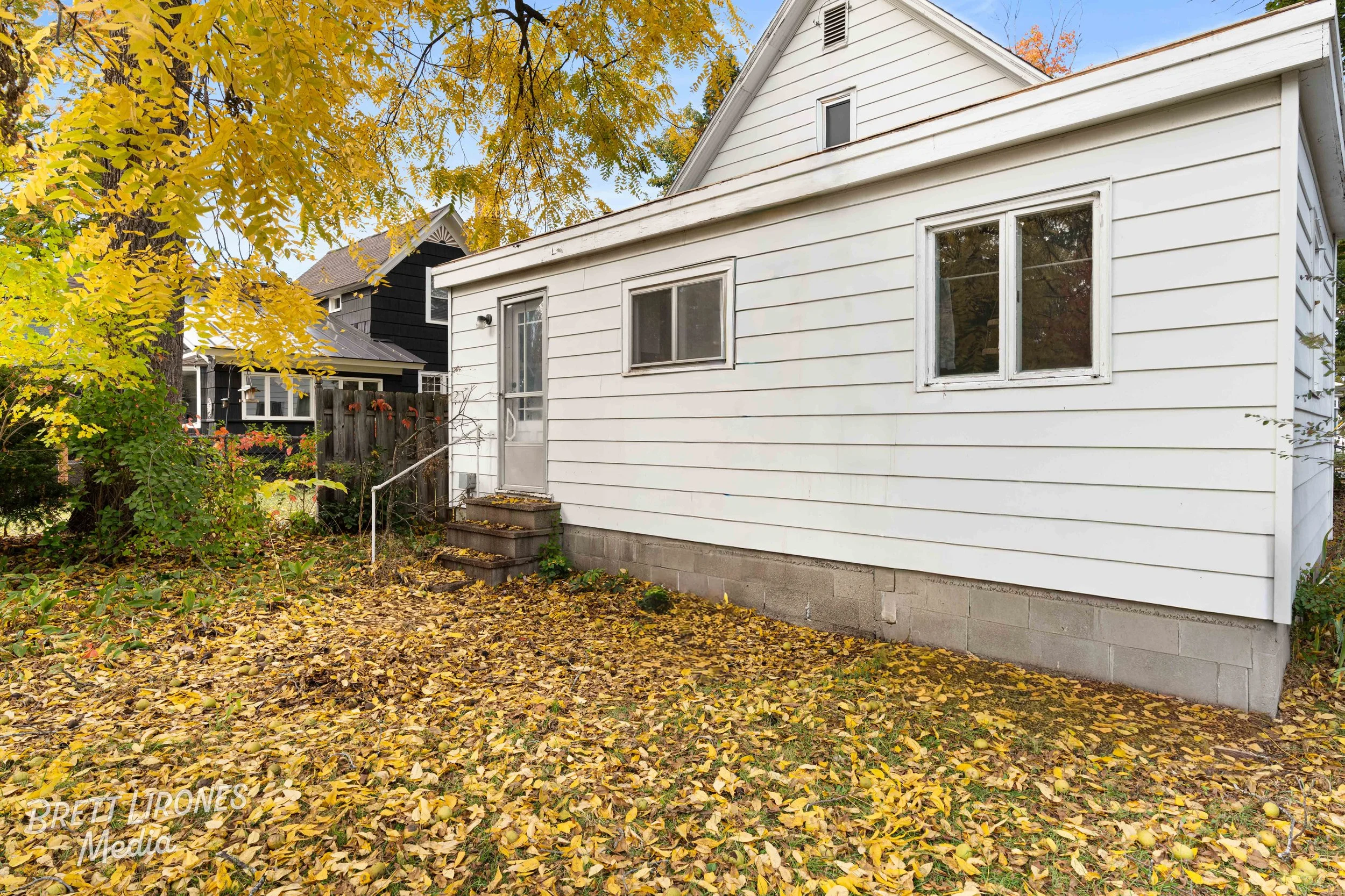 Backyard of a house in autumn with yellow fallen leaves on the ground, white siding, and trees with yellow leaves.