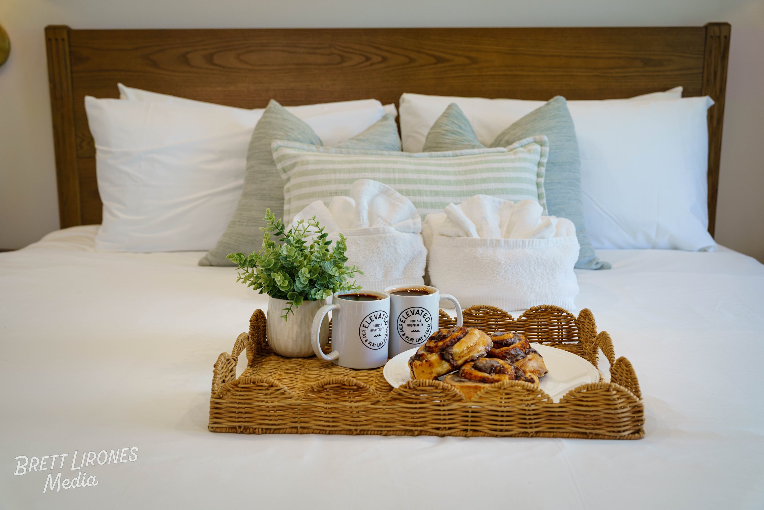 A wicker tray with two cups of coffee, a small potted plant, and a plate of cinnamon rolls placed on a white bed with pillows against a wooden headboard.