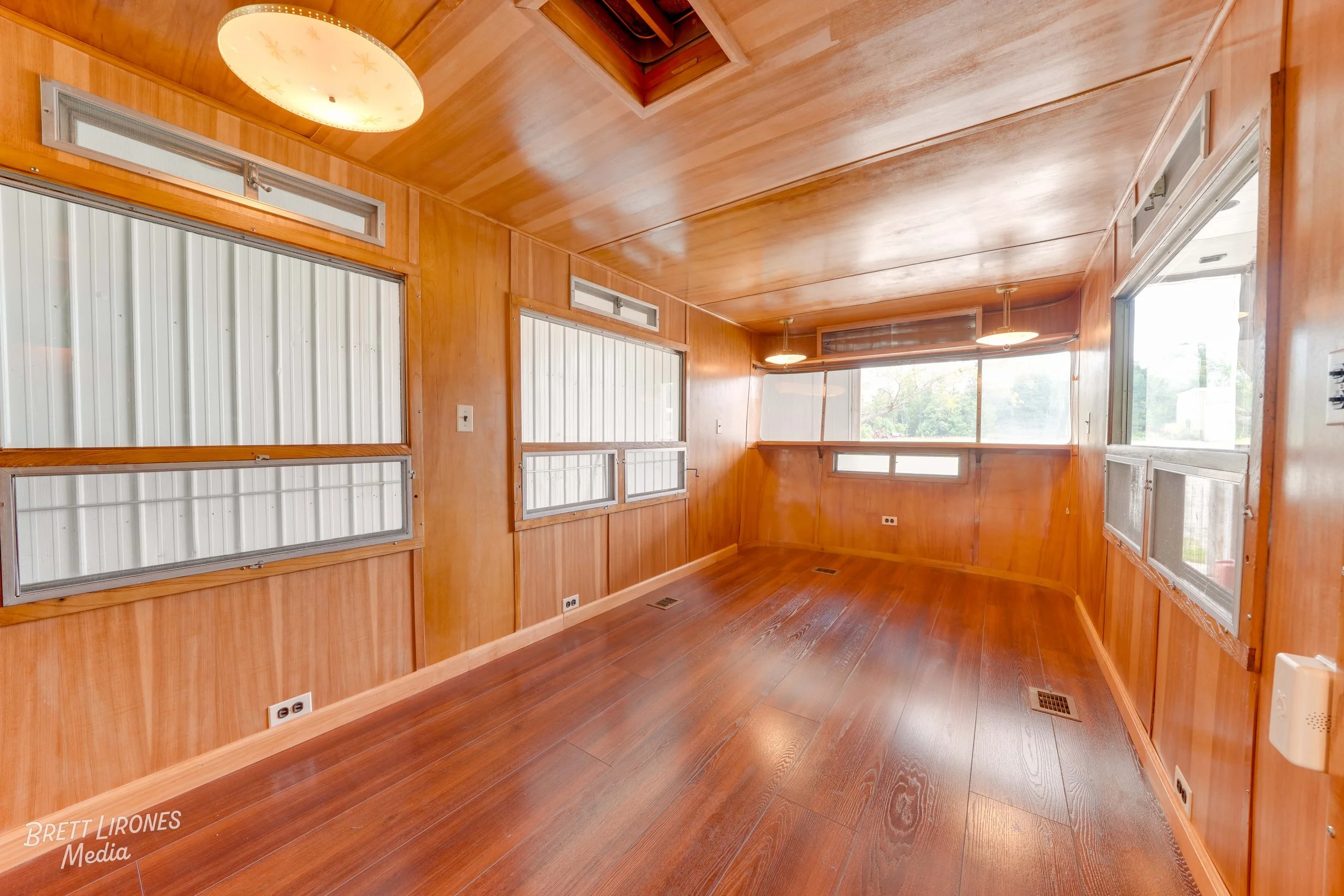 Empty room with wooden walls, ceiling, and floor, featuring multiple windows and ceiling lights.