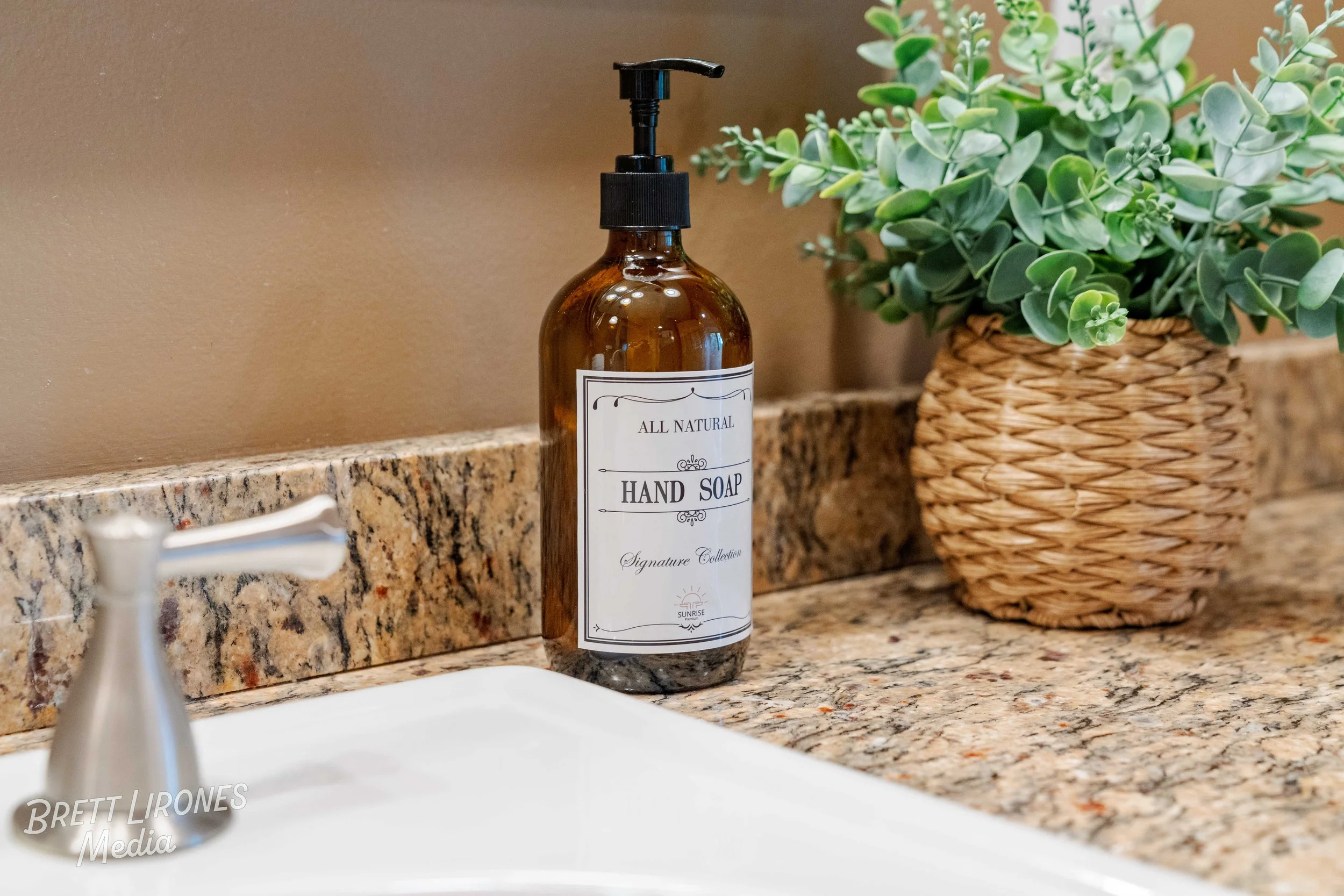 A brown glass bottle with a black pump labeled 'All Natural Hand Soap' on a granite bathroom countertop next to a woven basket with green artificial plant, partially visible faucet in foreground.