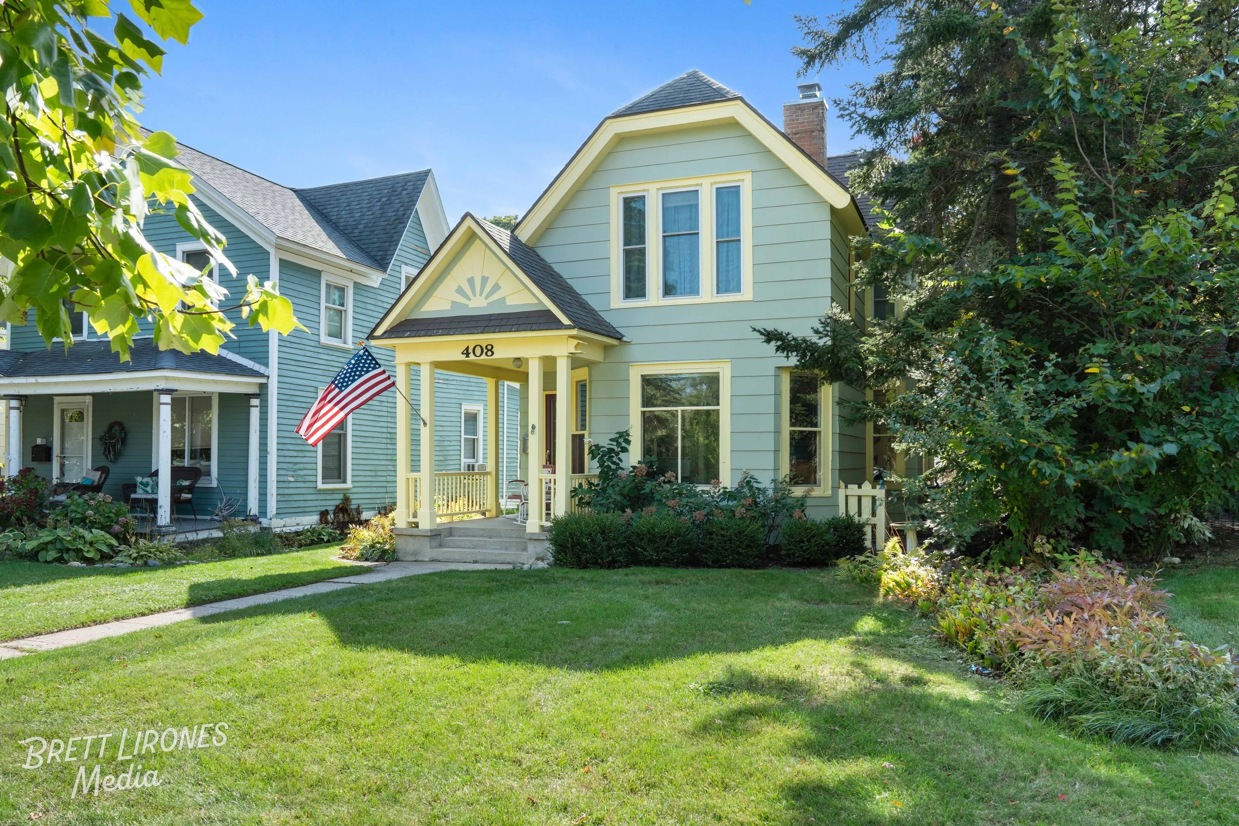 A light green, two-story house with the number 408 on the front porch, featuring a small set of stairs leading to the entrance, and an American flag mounted near the doorway. The house has yellow trim around the windows and porch, with lush green shr
