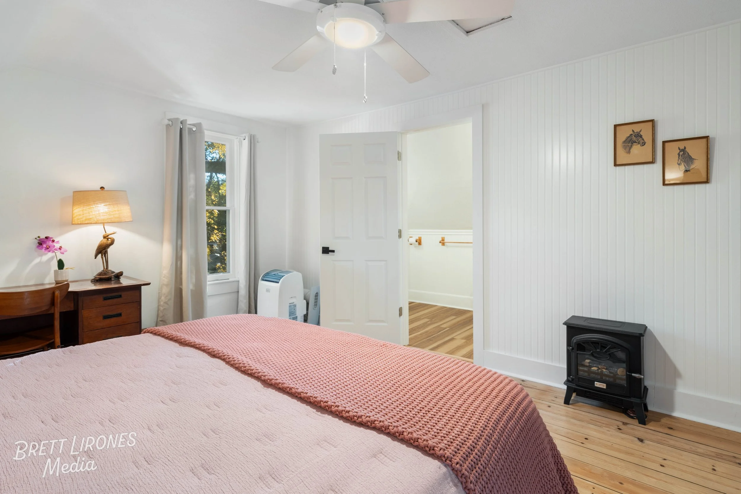 Interior of a bedroom with a bed covered in a pink blanket, a wooden nightstand with a lamp and decorative bird sculpture, a window with white curtains, an air conditioner, a partially open door leading to a hallway with hardwood flooring, and framed