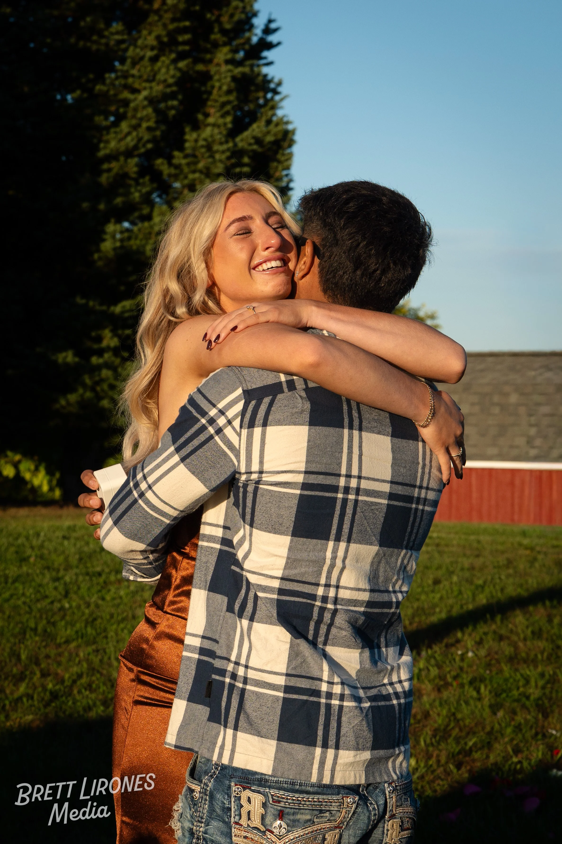 A man and a woman are hugging outdoors, with the woman smiling and eyes closed, during a sunny day.