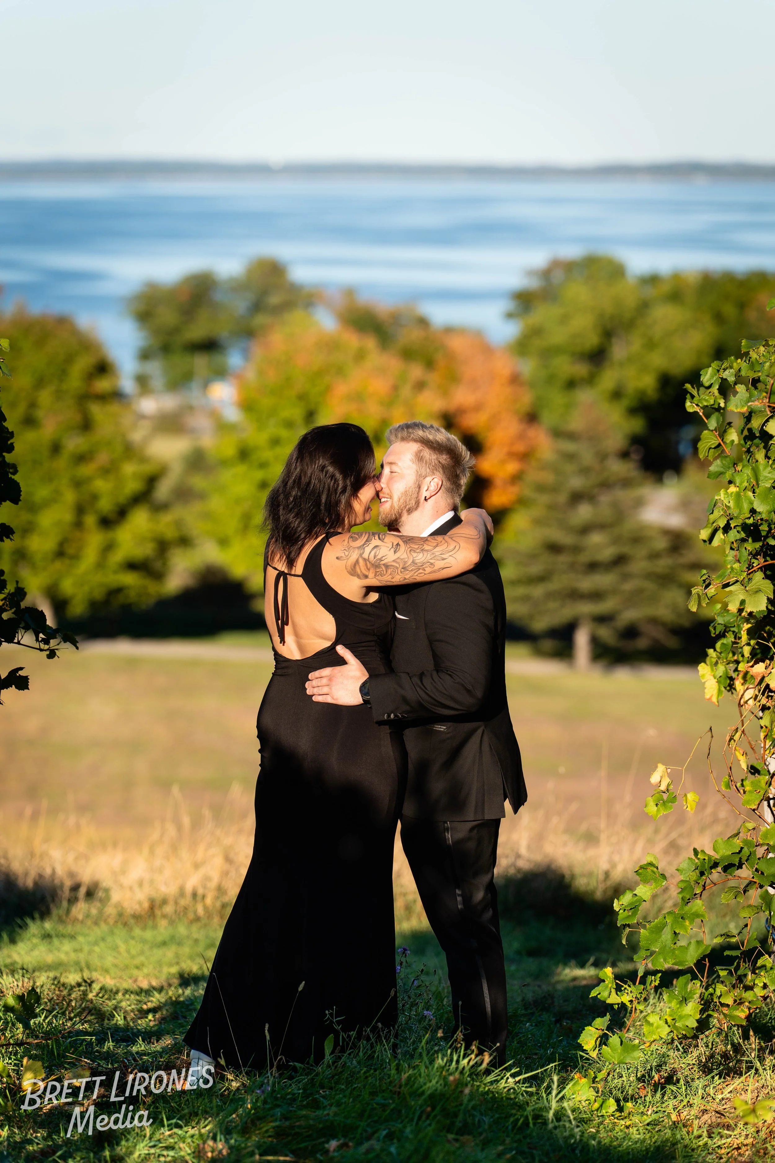 A couple dressed in formal attire embracing outdoors with trees and water in the background, during daytime.