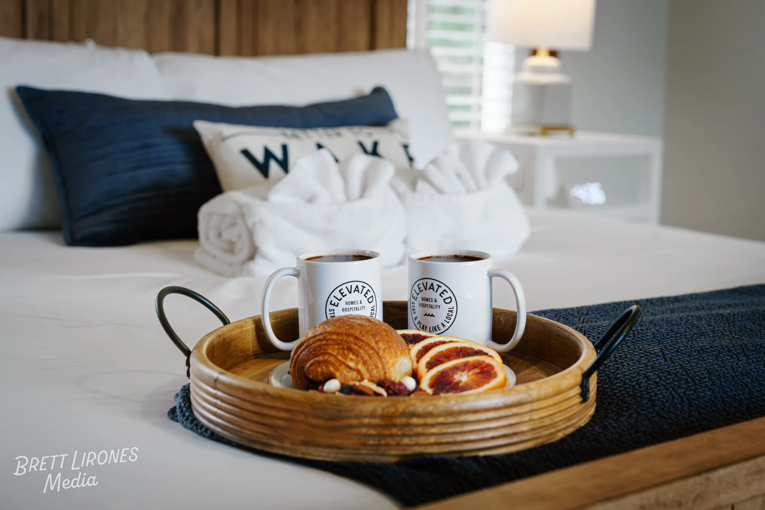 A breakfast tray with two mugs of coffee, croissants, and orange slices on a bed in a cozy bedroom with white bedding, navy pillows, and a wooden headboard.