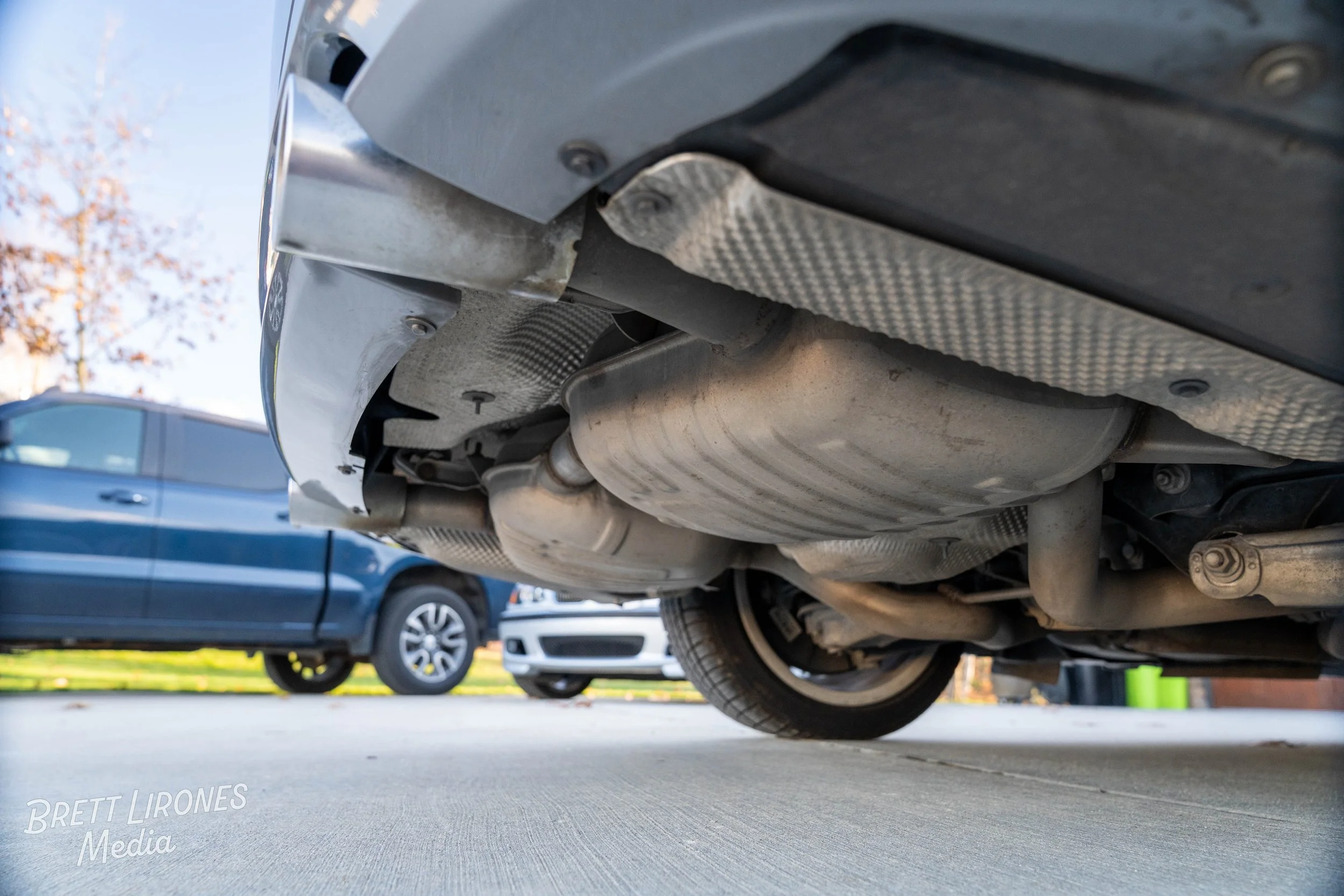 Underneath view of a car showing the exhaust system and muffler, with another vehicle parked in the background.
