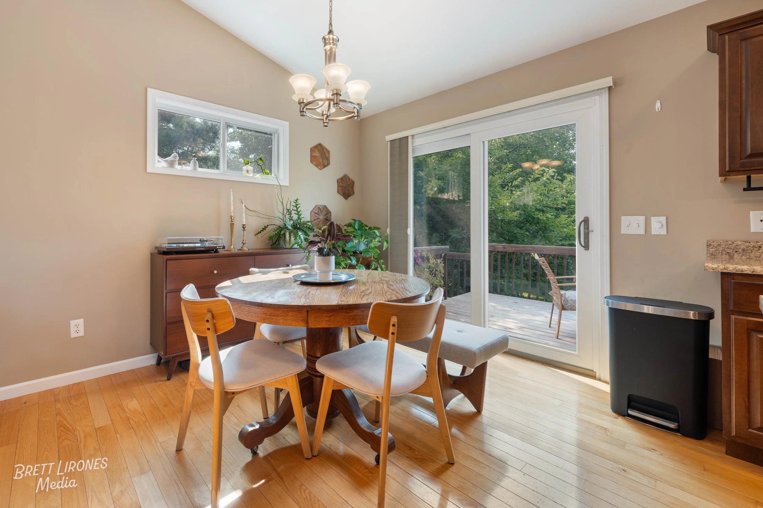 Kitchen dining area with a wooden round table, four modern chairs, a sideboard with plants, a chandelier, sliding glass door leading to a deck with outdoor chairs, and a small window.