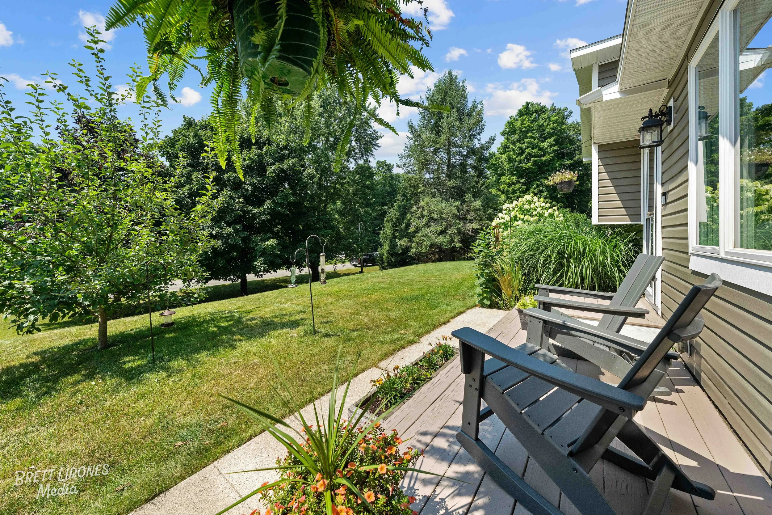 View of a backyard with a wooden deck, two gray Adirondack chairs, plants, a small tree, a well-maintained lawn, trees, and a street in the distance under a blue sky with clouds.