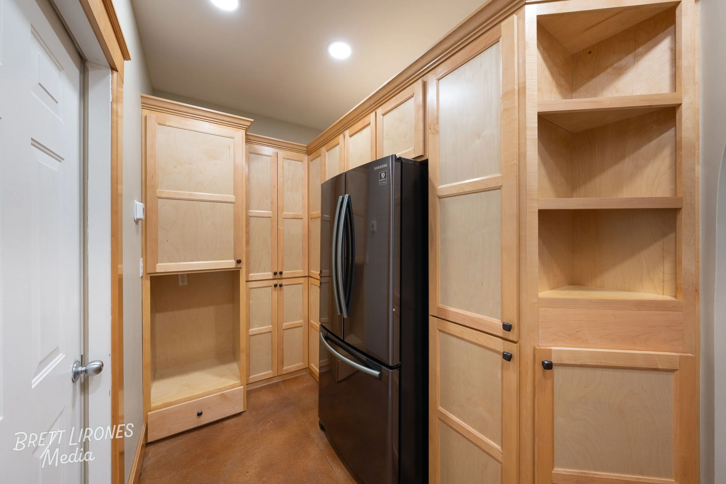 A laundry room with unfinished wooden cabinets surrounding a black Samsung refrigerator, with the cabinets empty and a brown floor.