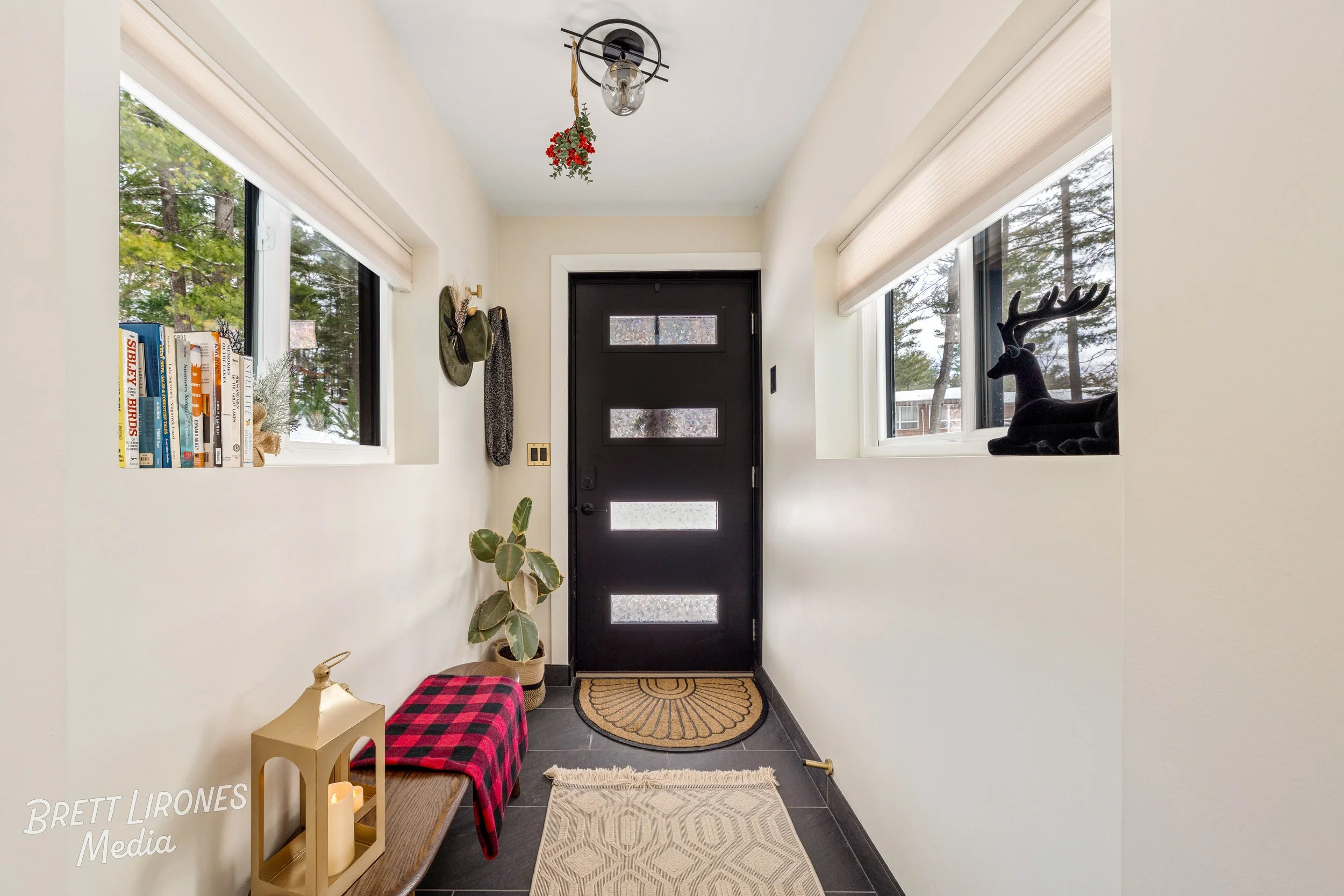Entryway with black front door, two windows, beige walls, black tile floor, decorative plants, books, and a black reindeer figurine on the window sill.