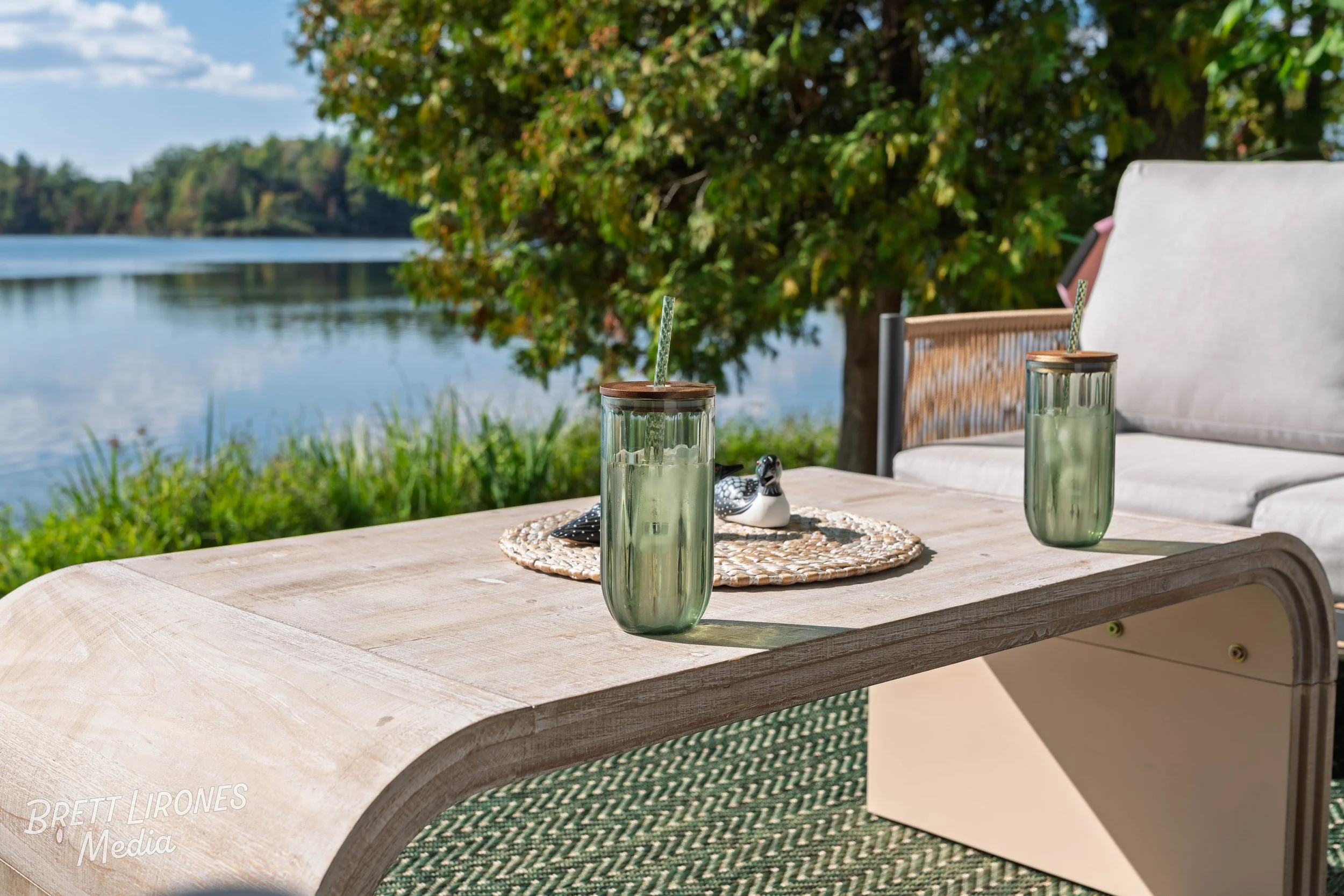 Outdoor patio table near a lake with two green glass jars with straws, a decorative tray, and a small ceramic duck figurine. A tree with green leaves and a blue sky are in the background.