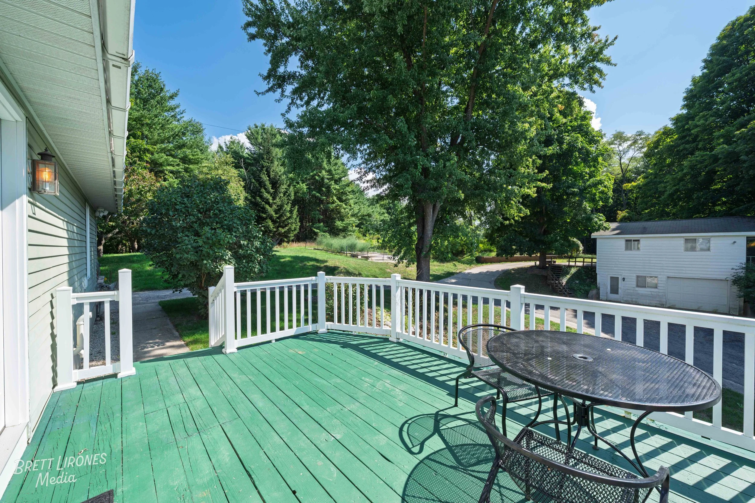 Green wooden deck with a black metal patio table and chairs, white railing, adjacent house with exterior lights, large trees, neighboring house, sunny day.