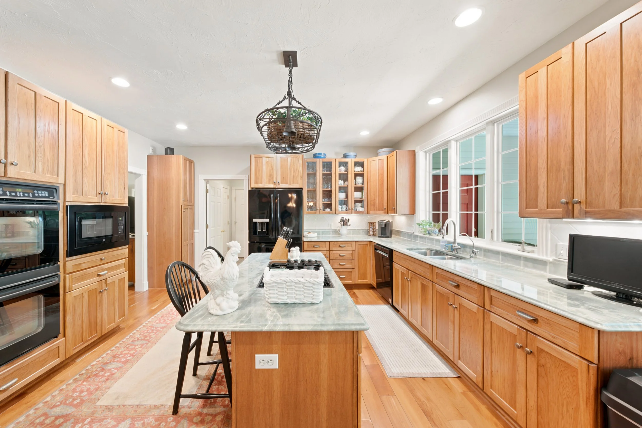 A spacious kitchen with light wooden cabinets, a large countertop, and a dining table. The kitchen features a black refrigerator, built-in microwave, and large windows letting in natural light. There is a hanging lantern-style light fixture above the