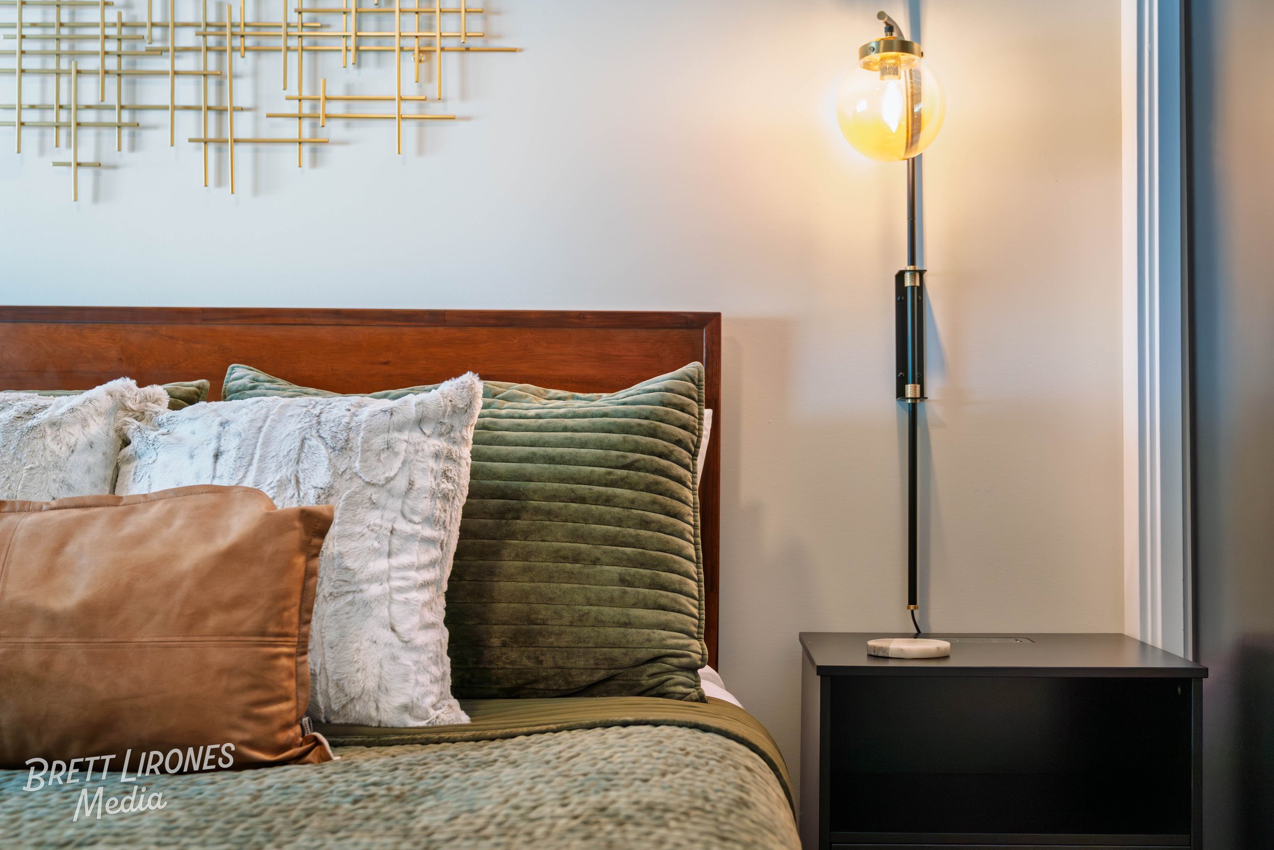 Close-up of a bedroom with pillows on a bed, a wooden headboard, a black nightstand, and a modern wall-mounted lamp with a large exposed bulb, white wall background, and geometric wall art.